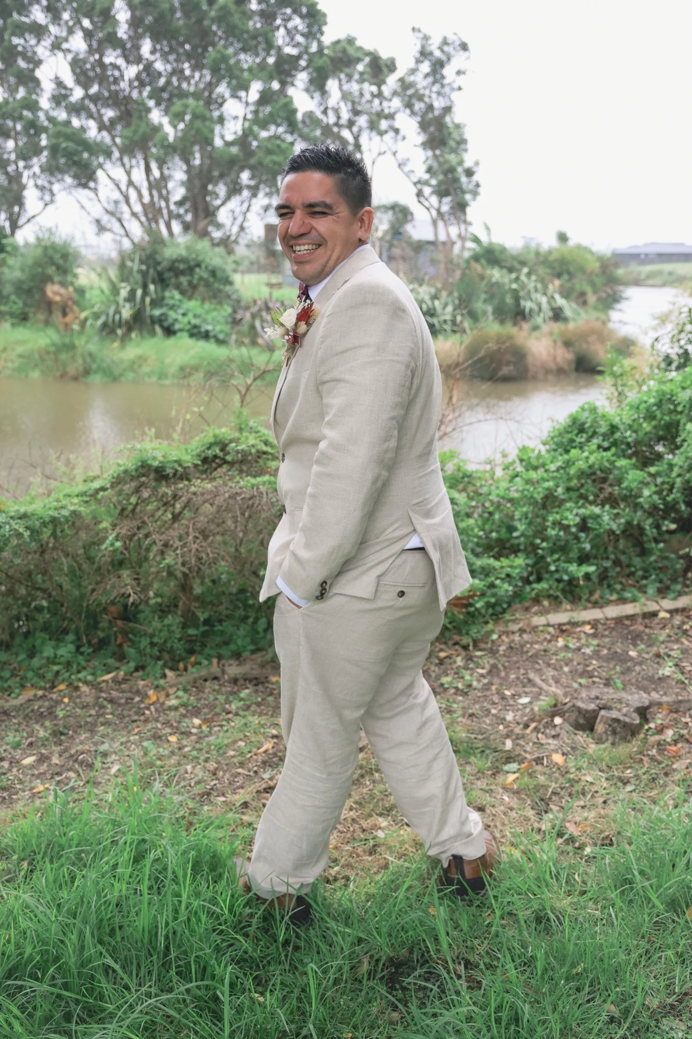 A man dressed in a light beige suit and dark shoes standing on a grassy area near a small body of water, smiling and looking away from the camera. The background features trees, shrubs, and a cloudy sky.