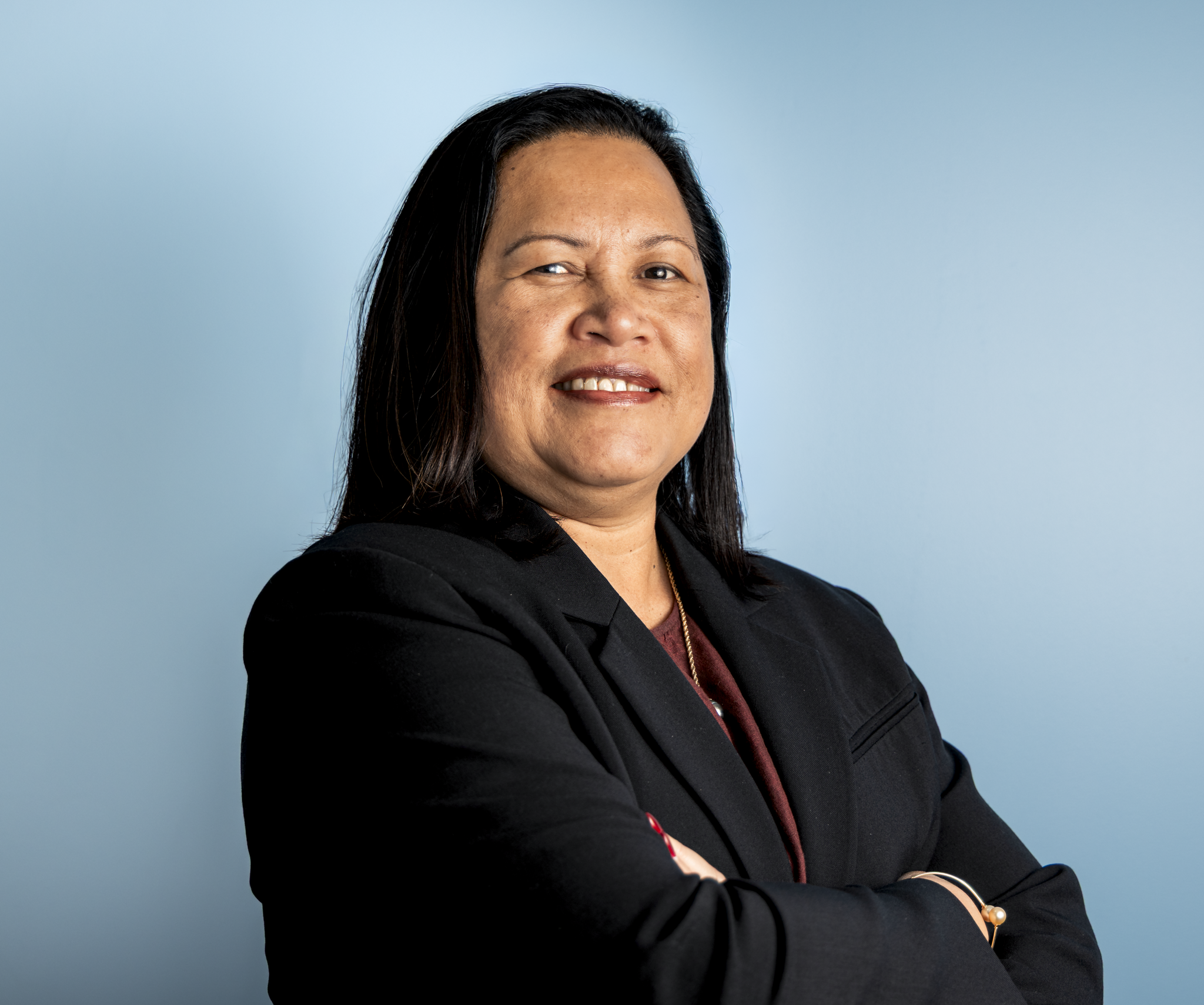 A woman with long black hair, wearing a black blazer and maroon shirt, standing against a blue background. She has a slight smile and is looking at the camera.