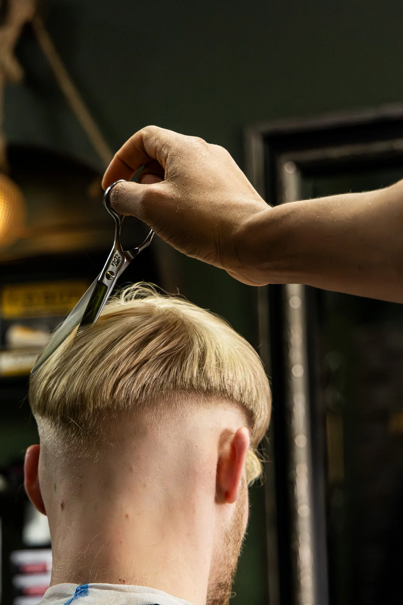 A person with short blonde hair getting a haircut, with a hairdresser using scissors on the hair. The person's face is not visible, focusing on the back of their head and the hairdresser's hand.