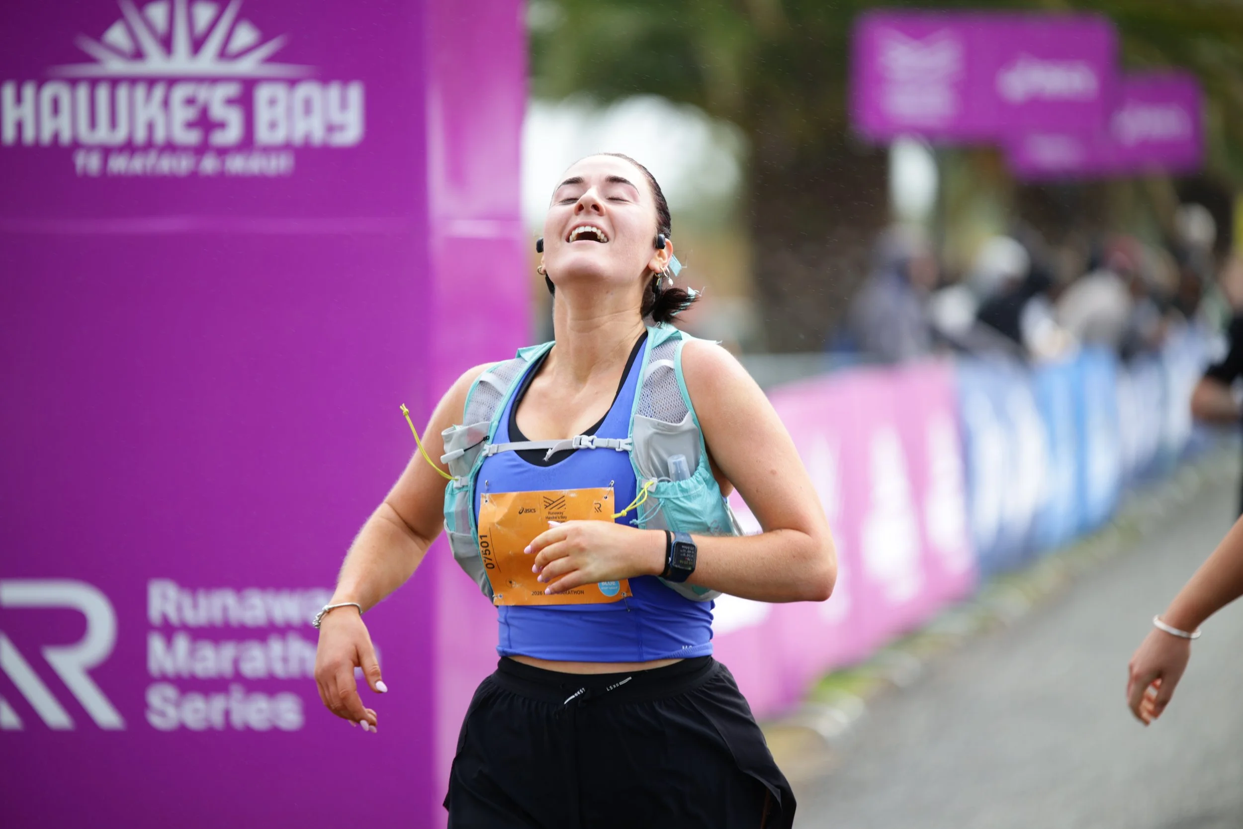 Female marathon runner smiling with eyes closed, wearing a gray and blue running vest, black shorts, and portable earbuds, running outdoors in front of a purple banner with white text at a race event.