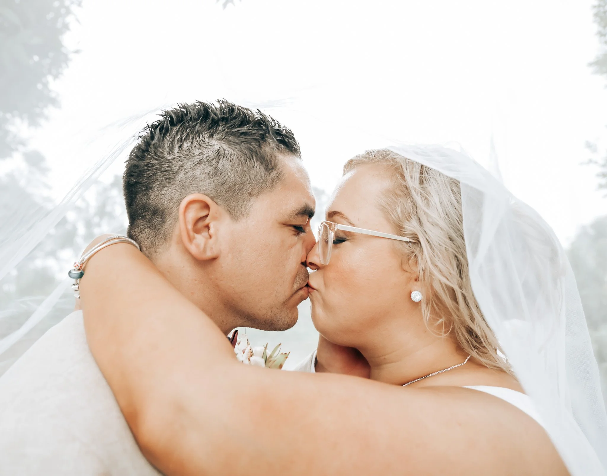 A couple about to kiss, with the woman's face visible through glasses and wearing a veil, and the man's face partly obscured, outdoors with trees in the background.
