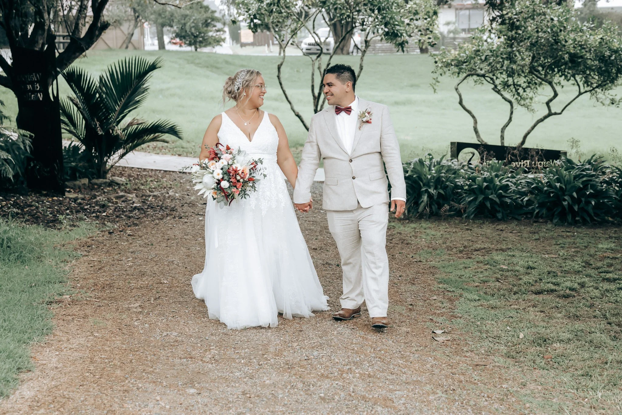 A bride and groom holding hands and smiling at each other during their outdoor wedding, surrounded by greenery and trees.