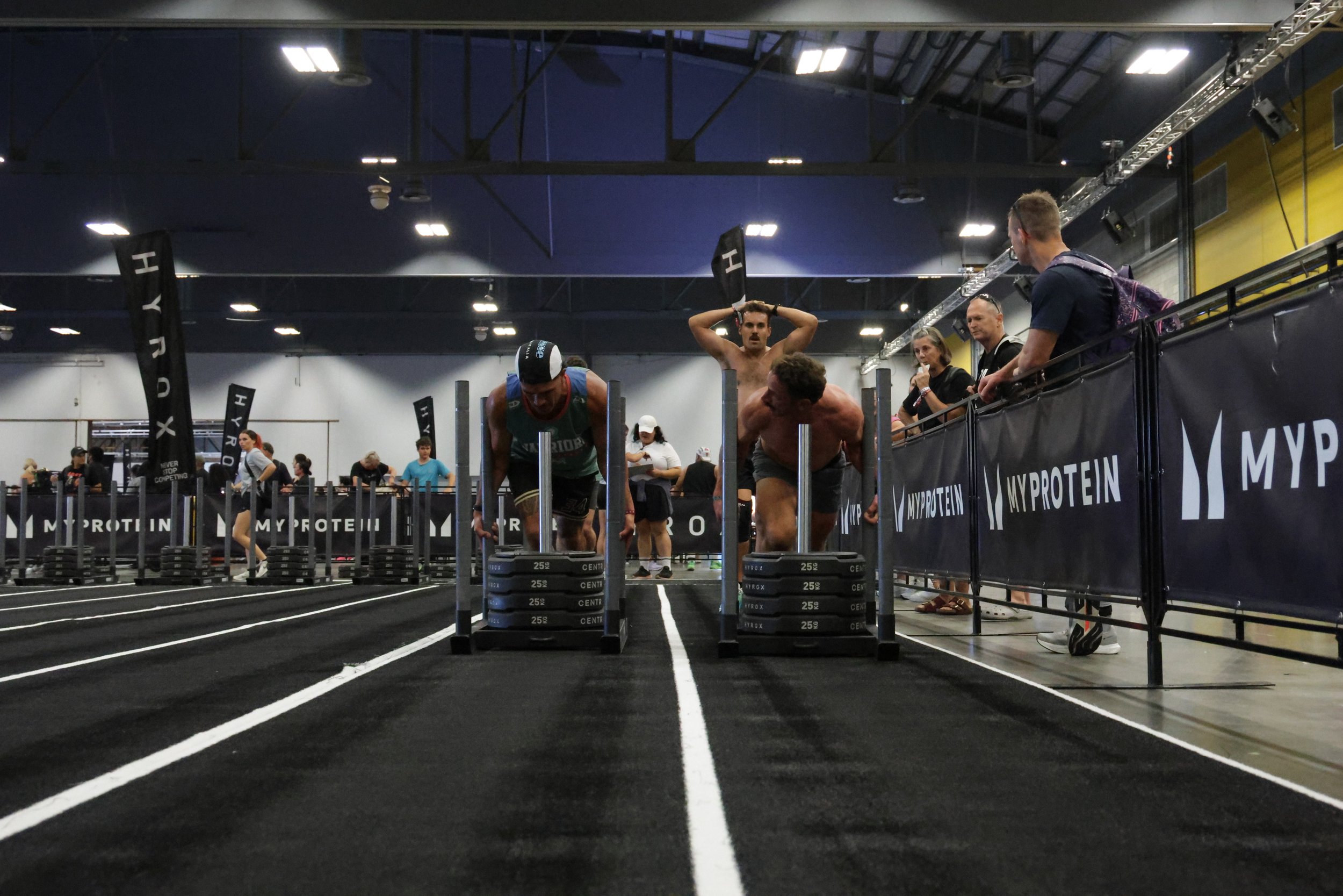 People participating in a fitness competition or workout in an indoor sports facility, with some lifting weights and others doing push-ups, surrounded by black barriers with branding.