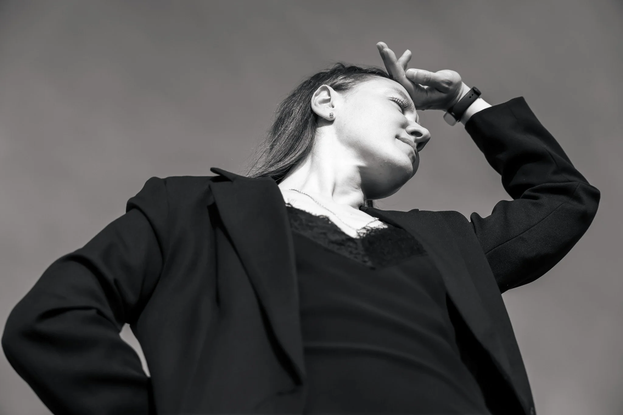 A woman in a black blazer with her eyes closed, smiling slightly, with her head tilted back and her hand raised above her forehead, against a plain background.
