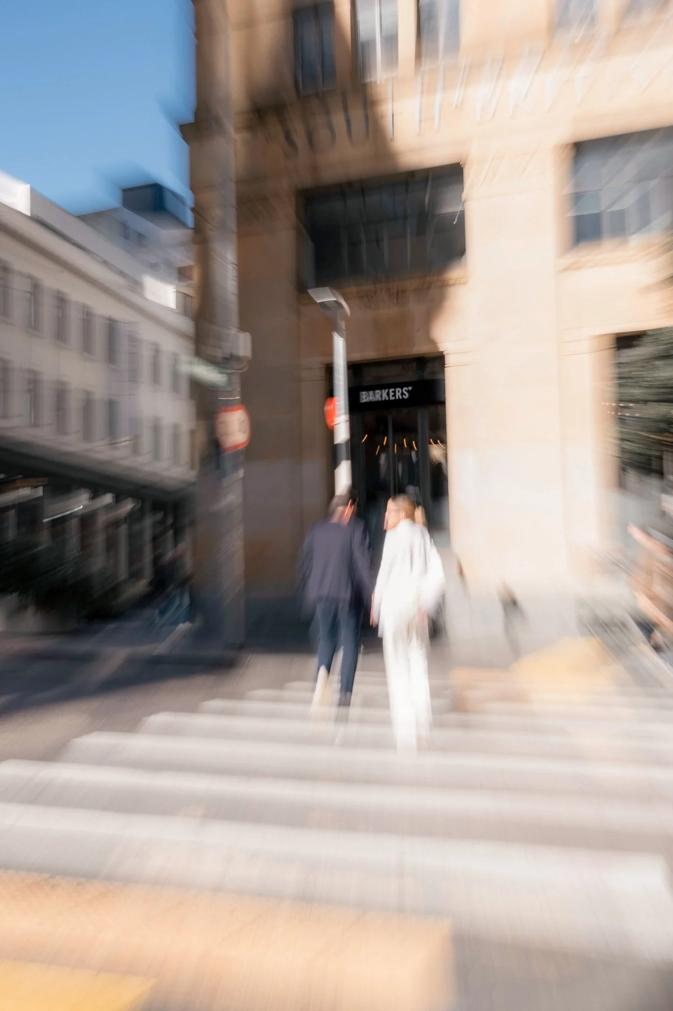 Two people walking across a crosswalk in front of a building with a store called 'BARKERS.' The photo appears blurry and is taken in an urban setting during daytime.