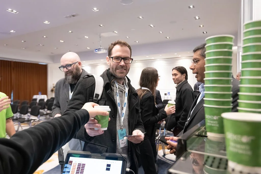 People socializing at a conference, some holding coffee cups, with a table of stacked green cups nearby.