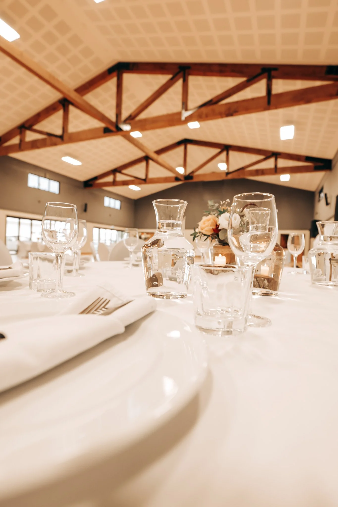 A table set for a formal event with water glasses, a small floral centerpiece, and a candle in a banquet hall with high wooden vaulted ceilings and skylights.