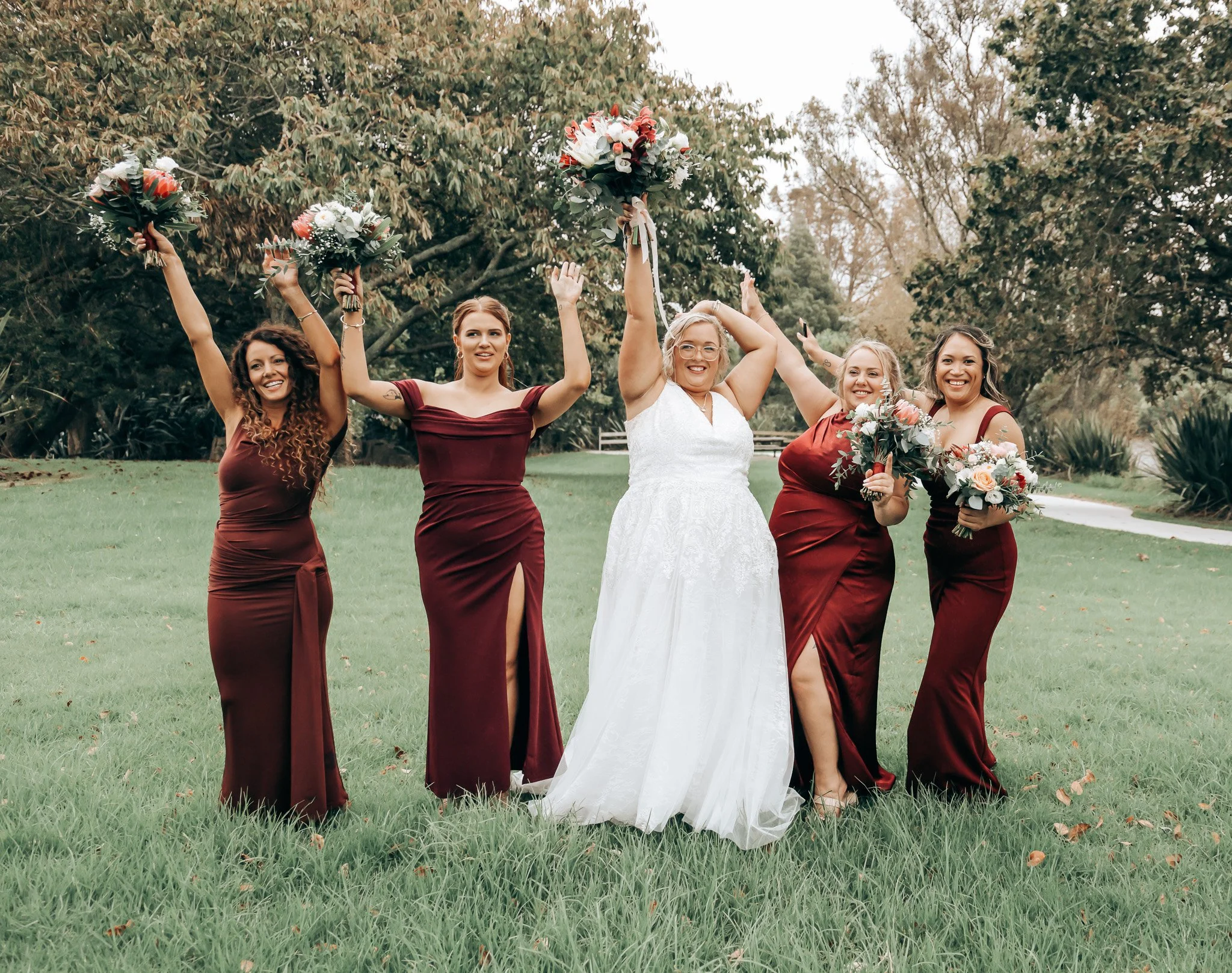 A bride and four bridesmaids celebrating outdoors, holding bouquets, with trees in the background.