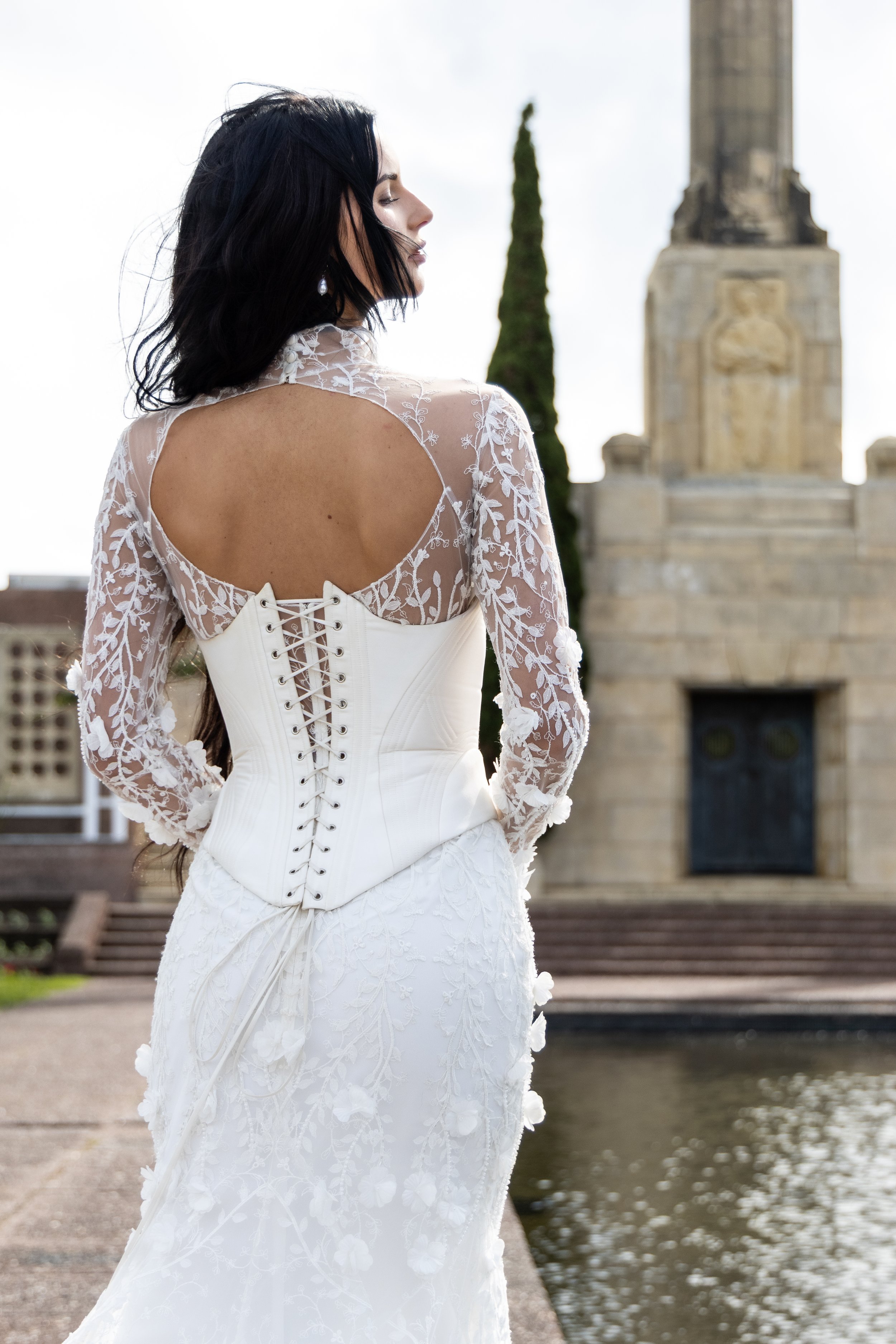 A woman in a white lace wedding dress with her back to the camera, standing outdoors near a body of water with a stone monument or building in the background.