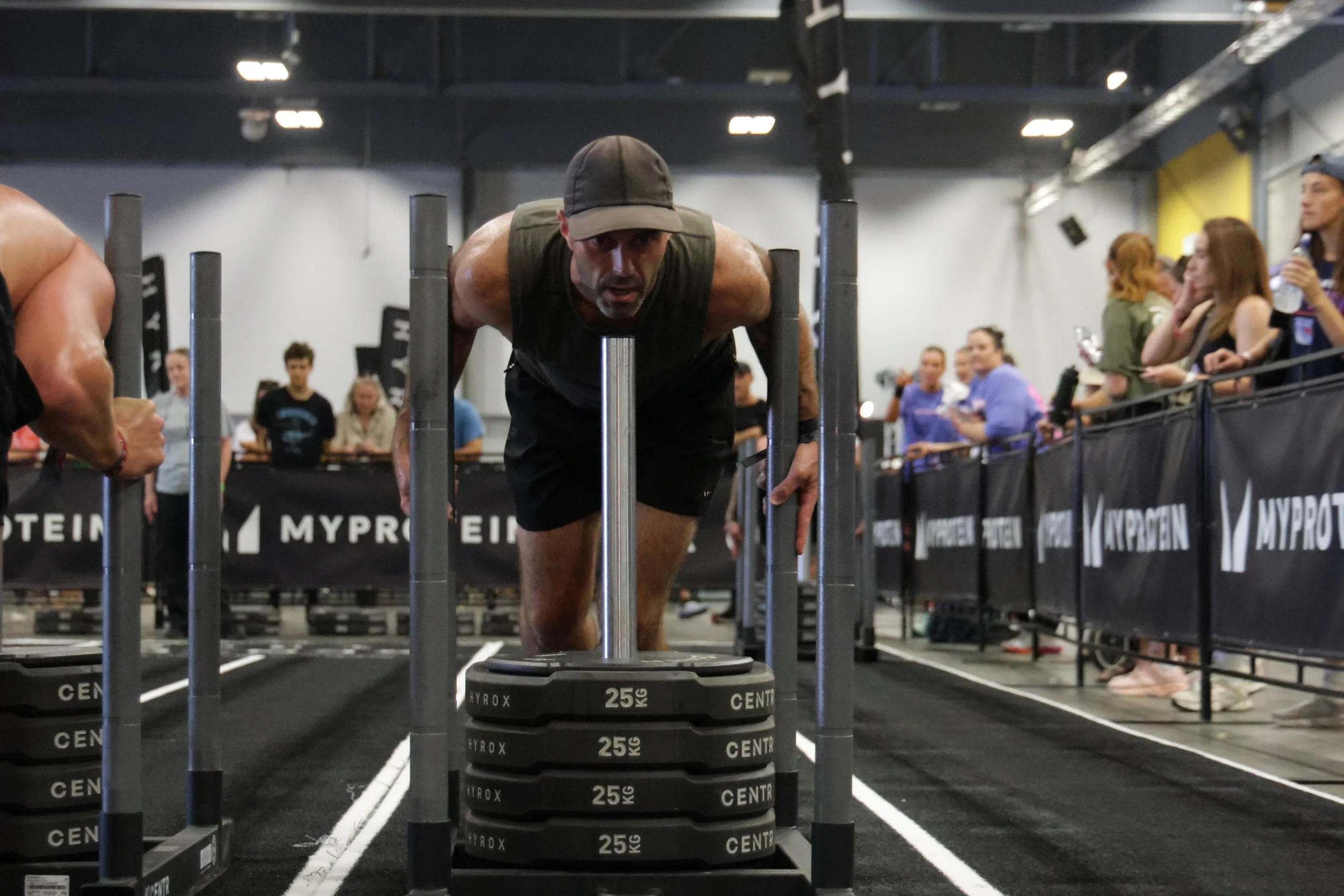 A man in a black cap and sleeveless shirt doing a push-up in an indoor gym, with weightlifting equipment and people in the background.