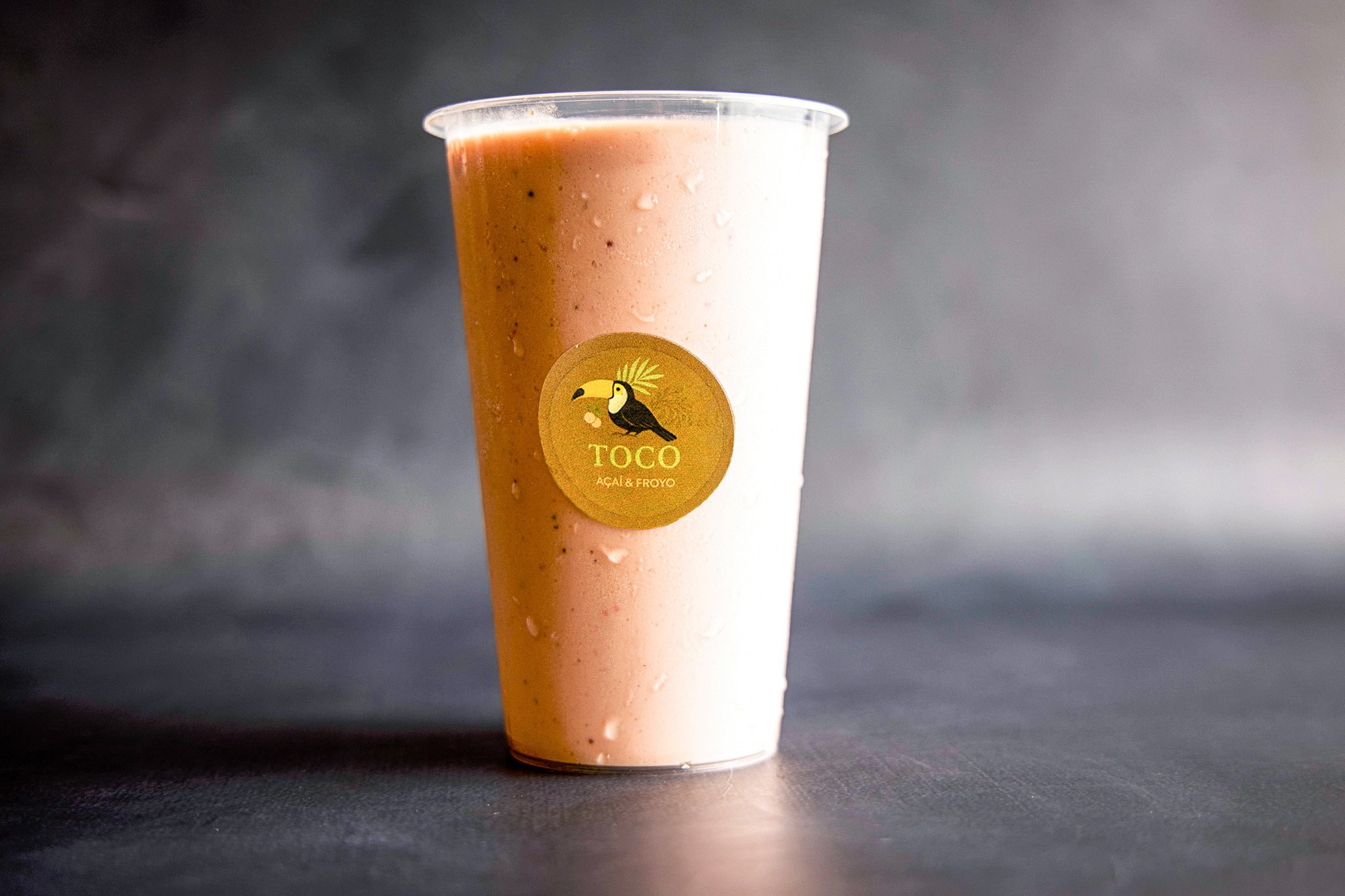 A plastic cup filled with pinkish smoothie, drops of condensation on the inside, featuring a round yellow logo with a toucan and the text 'TOCO Açaí & Froyo', placed on a dark surface with a plain gray background.