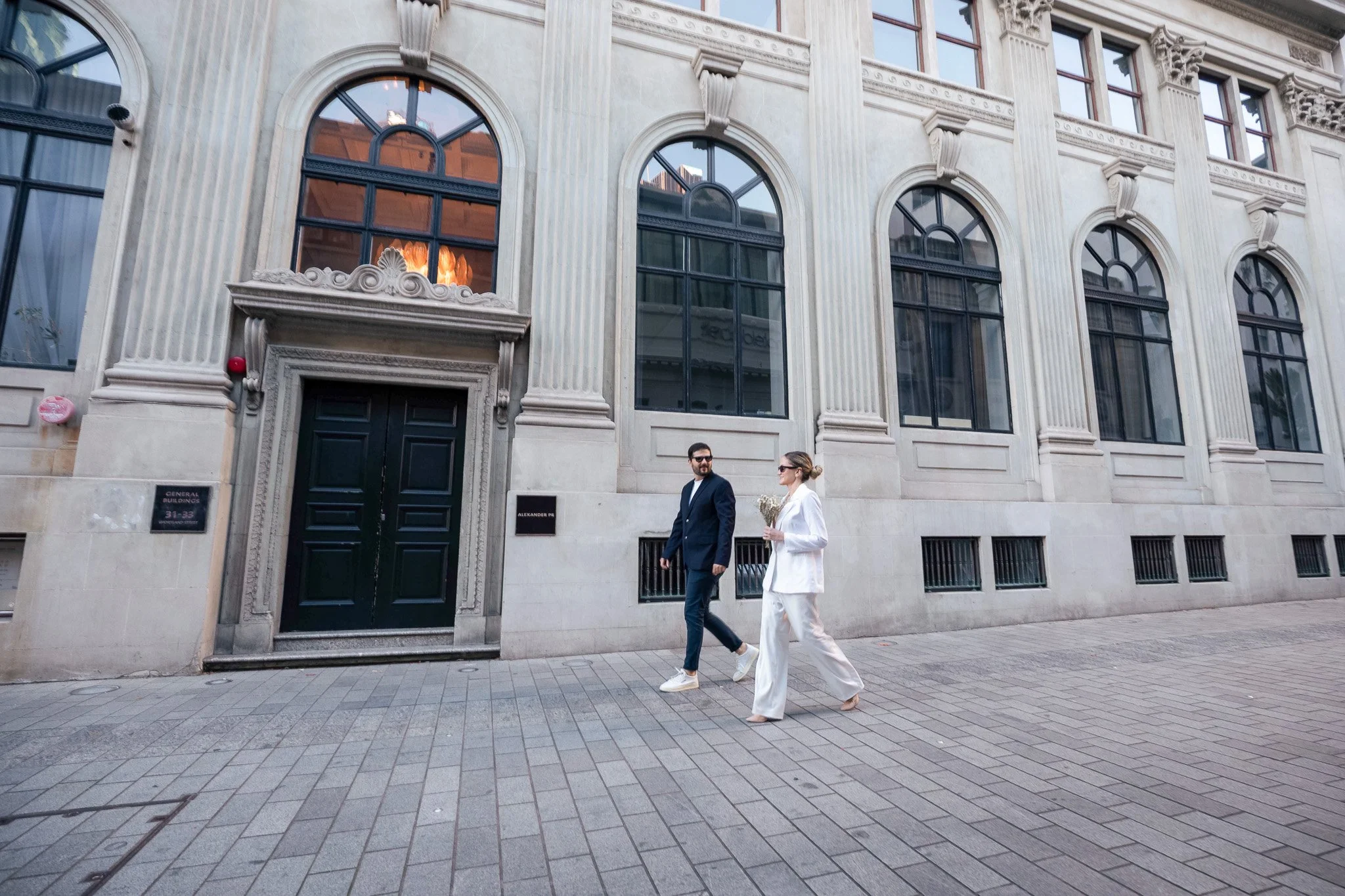 Two people walking past a historic building with large arched windows and decorative stonework. One person is dressed in a dark suit and sunglasses, and the other is in a white outfit carrying flowers.