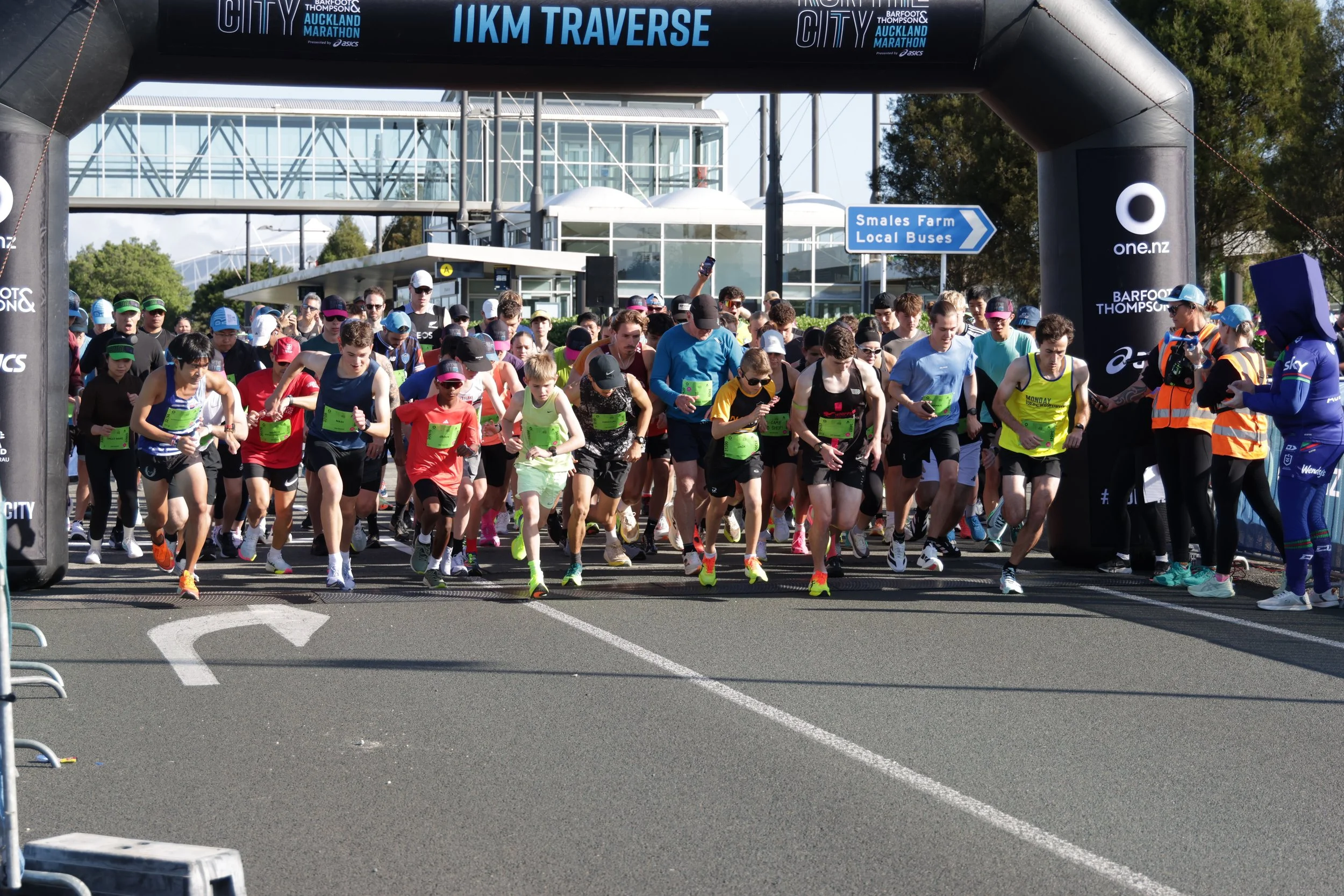 A large group of runners starting a race at the city marathon, visible under a black archway with blue digital signs indicating a 11 km traverse. The scene takes place outdoors near a modern, glass-fronted building, with spectators and event official
