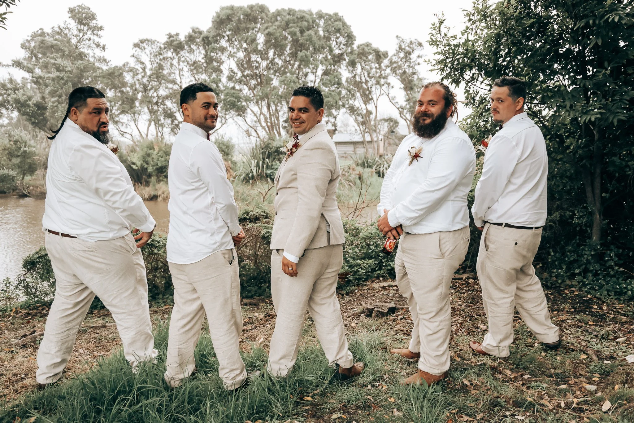 A group of six men dressed in wedding attire standing outdoors near a river, surrounded by trees and greenery.