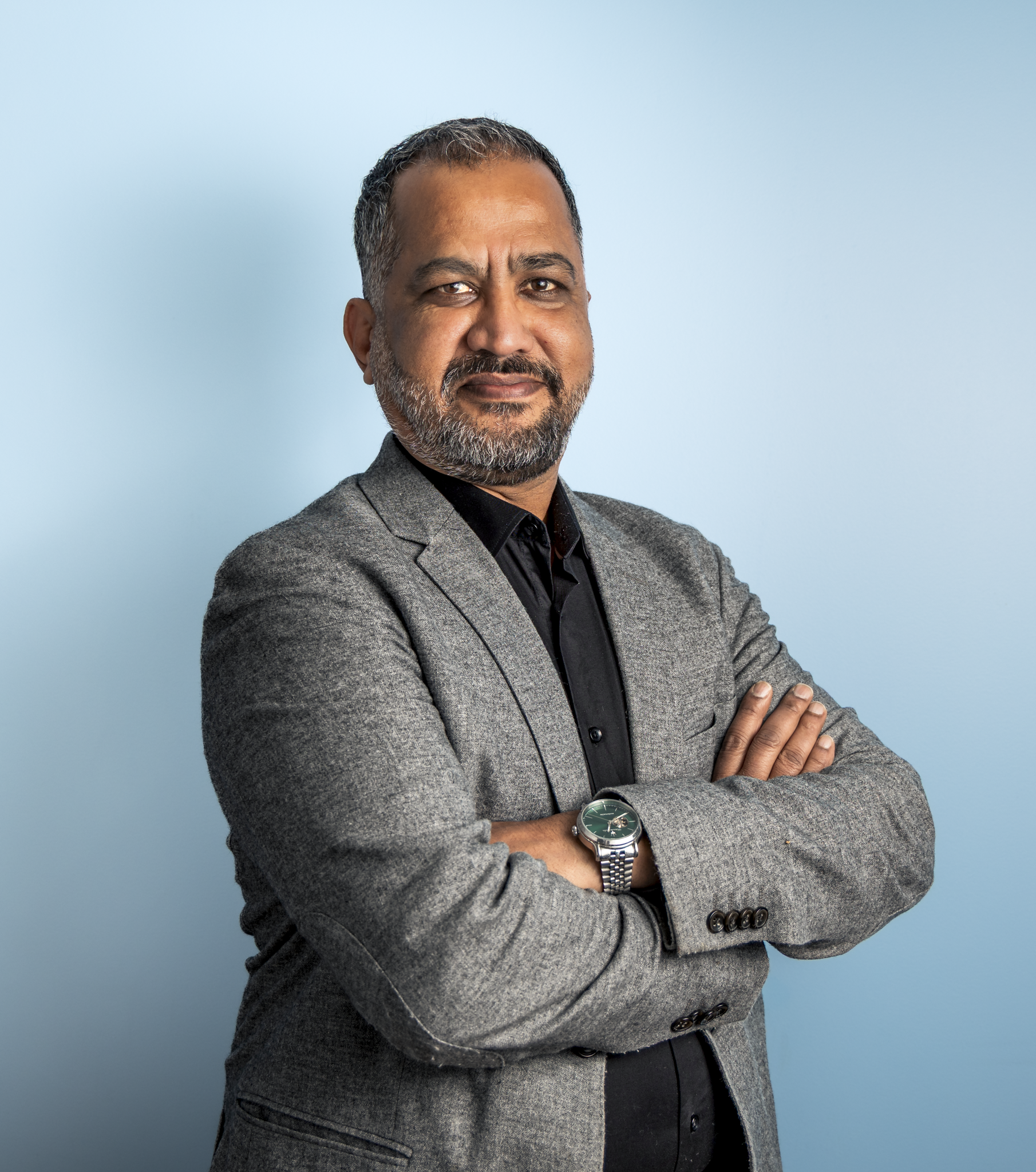 A confident middle-aged man with a beard and short hair, wearing a gray blazer, black shirt, and a wristwatch, stands with his arms crossed against a light blue background.