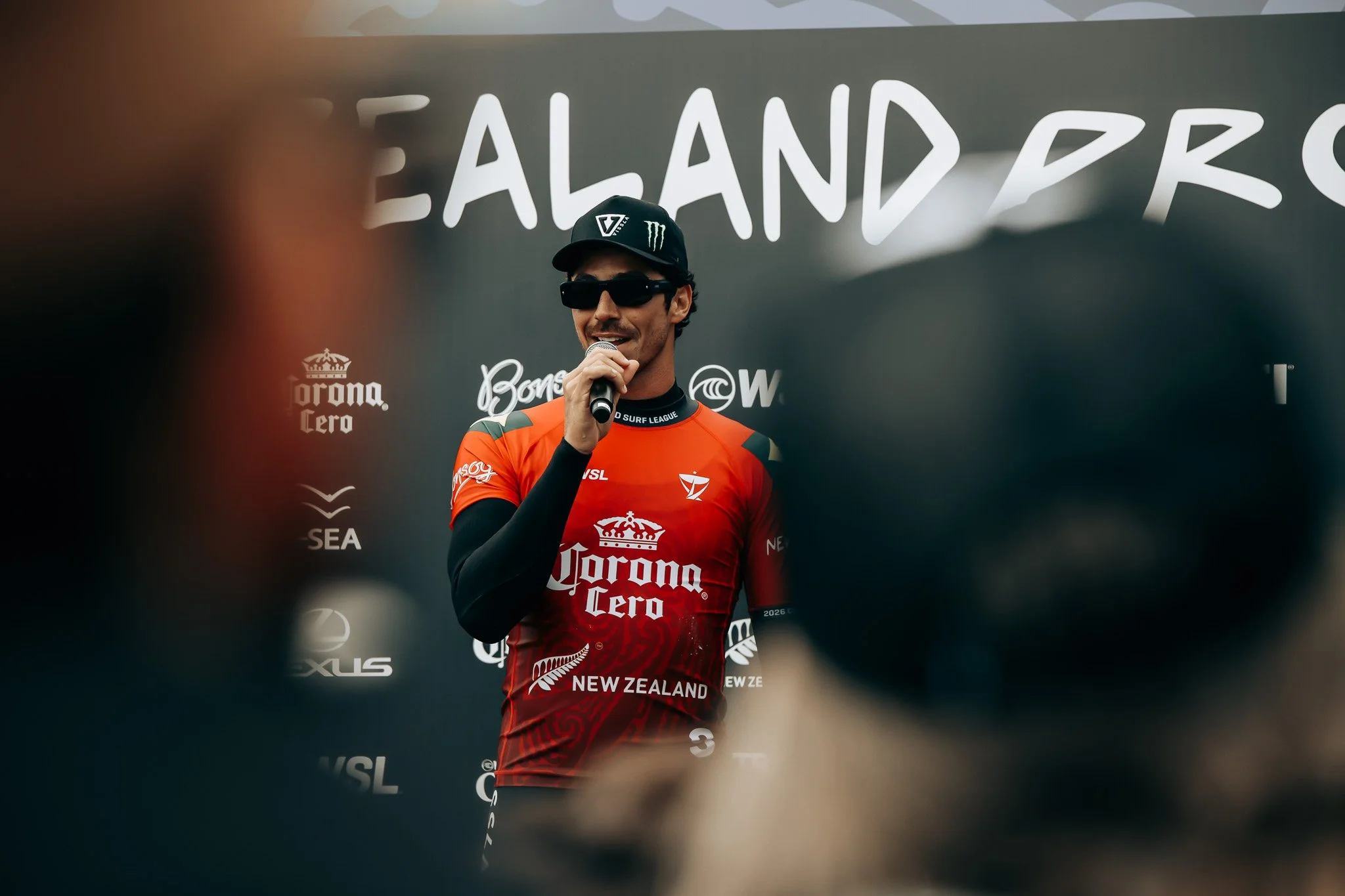 A swimmer in a red racing suit with New Zealand and sponsors’ logos, wearing sunglasses and a cap with a Monster Energy logo, standing in front of a mural with the word 'ISLAND' visible.