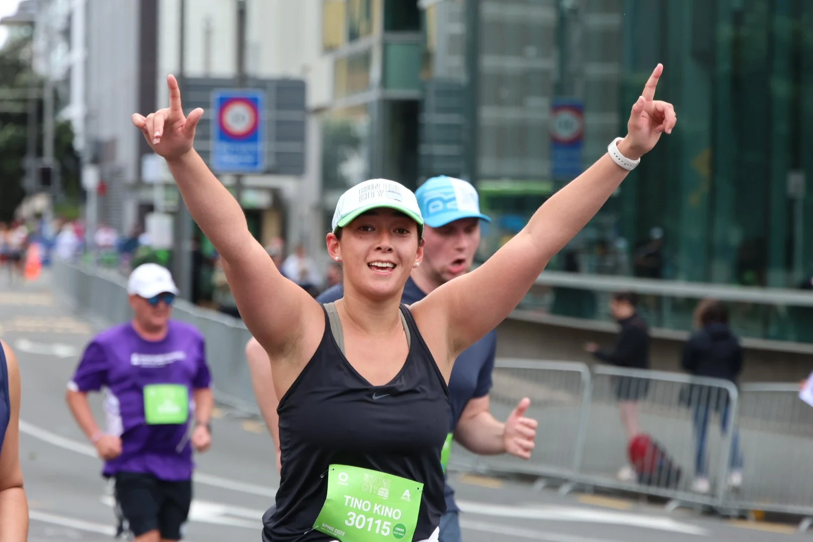 Female marathon runner smiling and raising her arms as she crosses the finish line during a race, with other runners and urban buildings in the background.