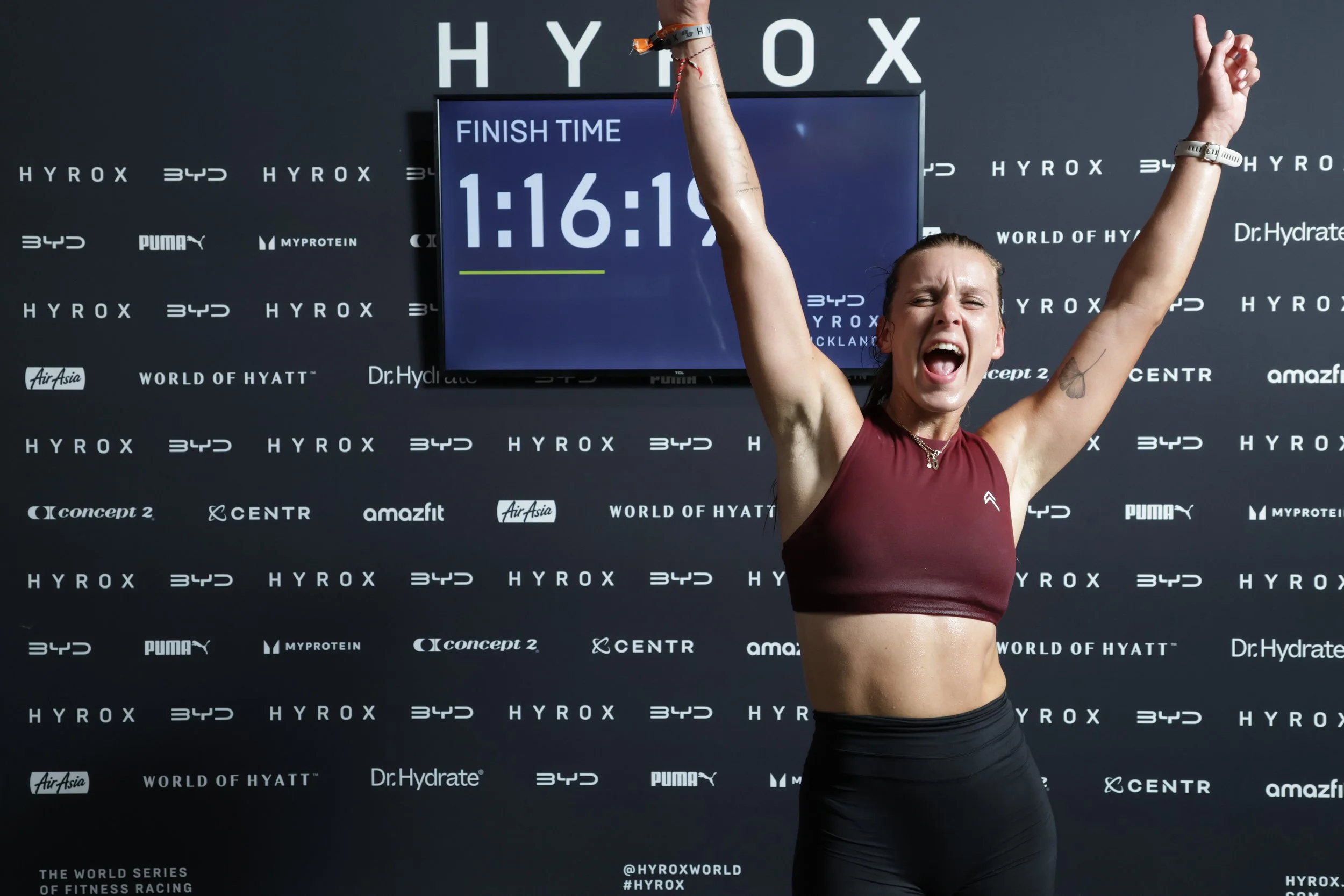 A woman with her arms raised in celebration, standing in front of a backdrop with various sponsor logos, and a large digital timer showing 1 hour, 16 minutes, and 19 seconds, indicating she finished a race.