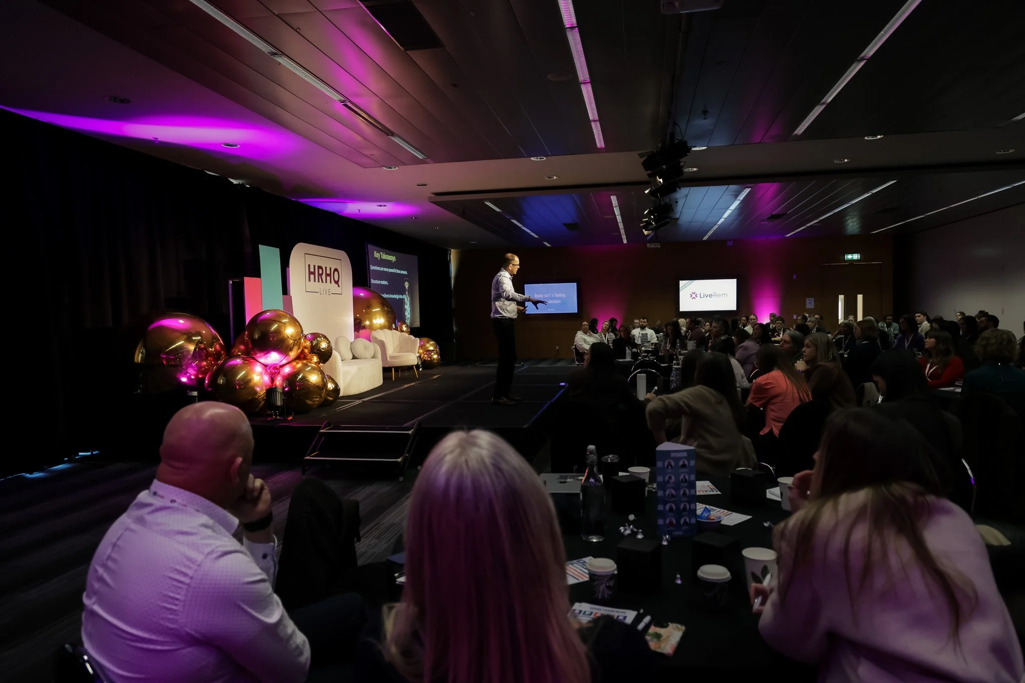 A conference or seminar with a speaker on stage and an audience seated at tables. The stage has a large sign with 'HRHQ Live' and decorative golden balloons. Purple lighting highlights the room.