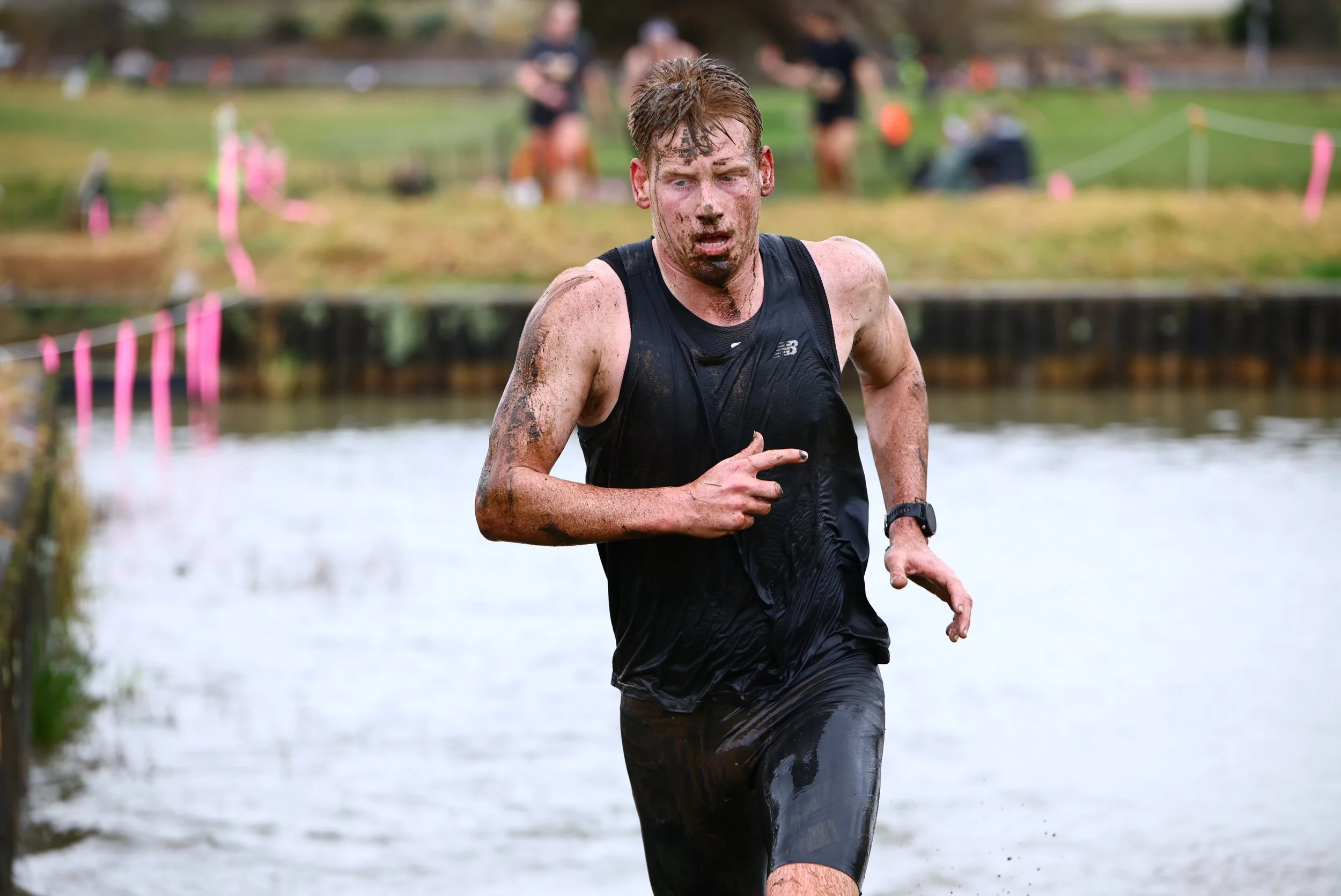 A man running through water during a muddy obstacle race, covered in mud and wearing a black athletic shirt.