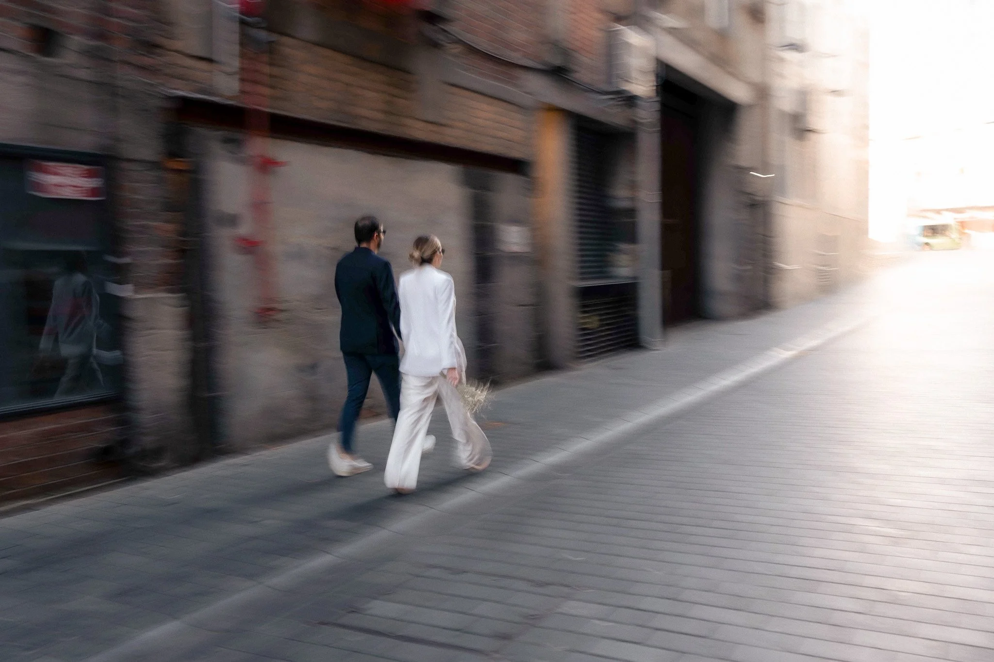 A man and woman walk together on a city sidewalk—she wears a white suit and he in a dark suit—with brick and concrete buildings in the background.