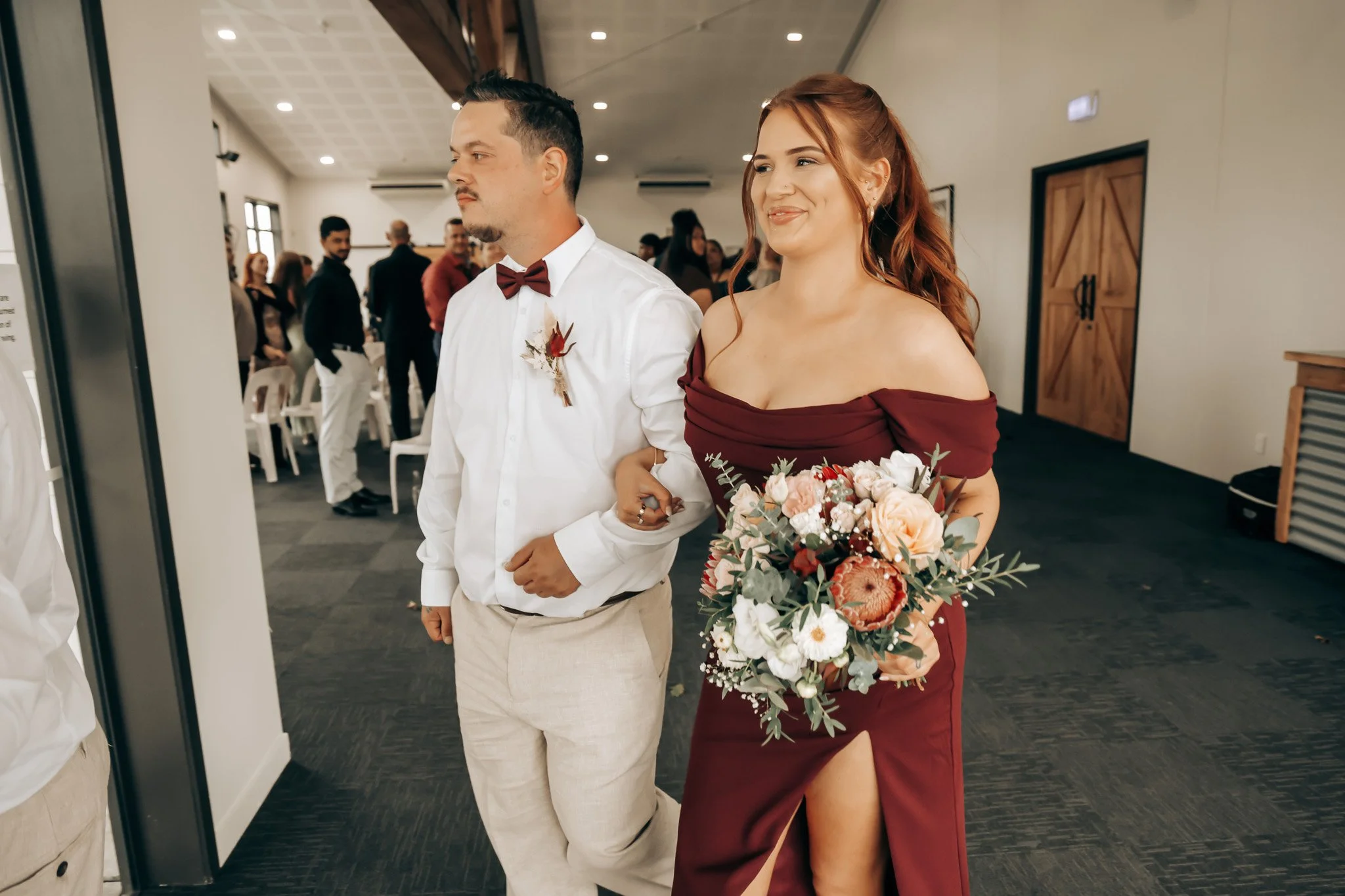 A young man and woman at a formal event, dressed elegantly. The woman is holding a large bouquet of flowers and smiling, while the man stands beside her with a serious expression. The room is filled with other people, some dressed in formal attire, a