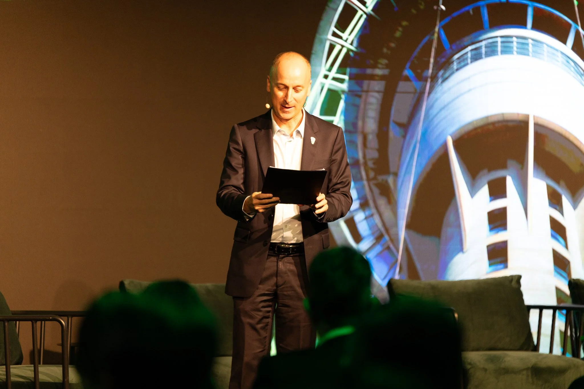 A man in a suit standing on stage, holding a tablet, with a brightly lit Ferris wheel behind him during a presentation or event.
