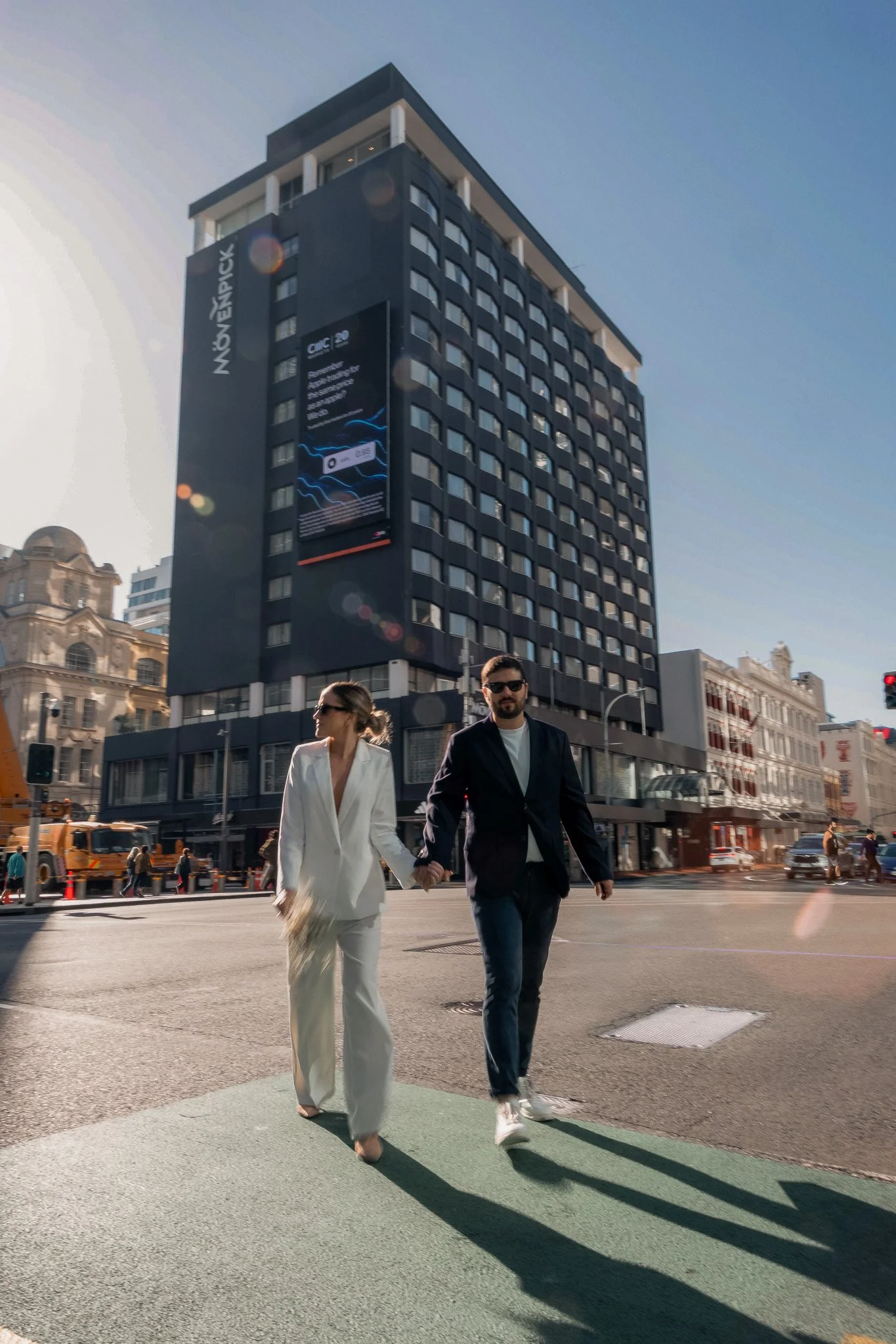 A couple holding hands and walking across a city street in the late afternoon, with tall buildings and a digital billboard in the background.