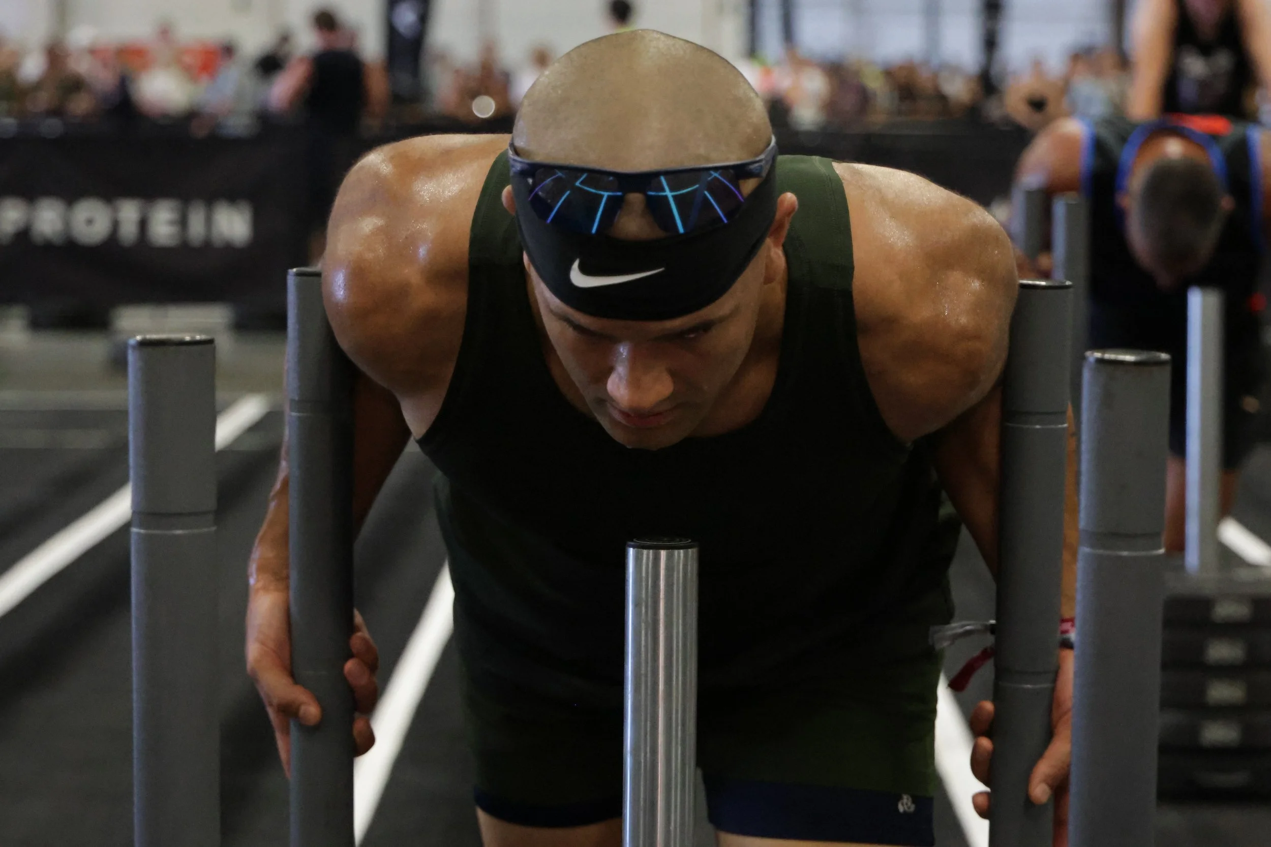 A man with a shaved head wearing sunglasses and a face mask, pushing a sled in a gym with other people in the background.