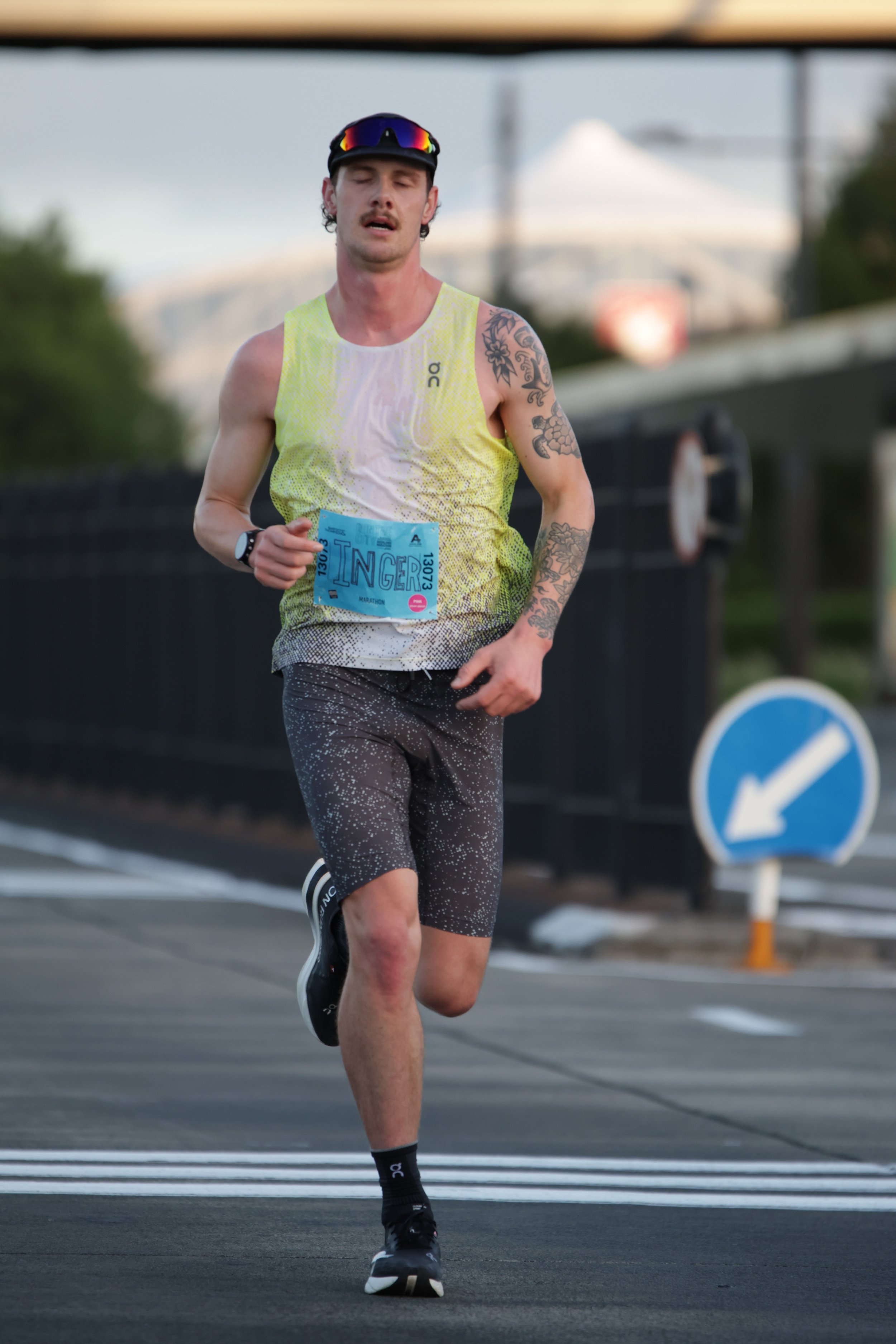 A male marathon runner wearing a yellow vest, black shorts, and black running shoes, holding a race bib numbered 13073, running outdoors on a paved road with traffic signs and a blurred vehicle in the background.