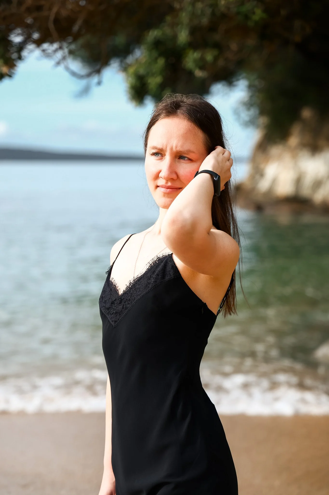 A young woman with long brown hair and light skin standing on a beach, wearing a black dress with lace details, with trees and a body of water in the background.