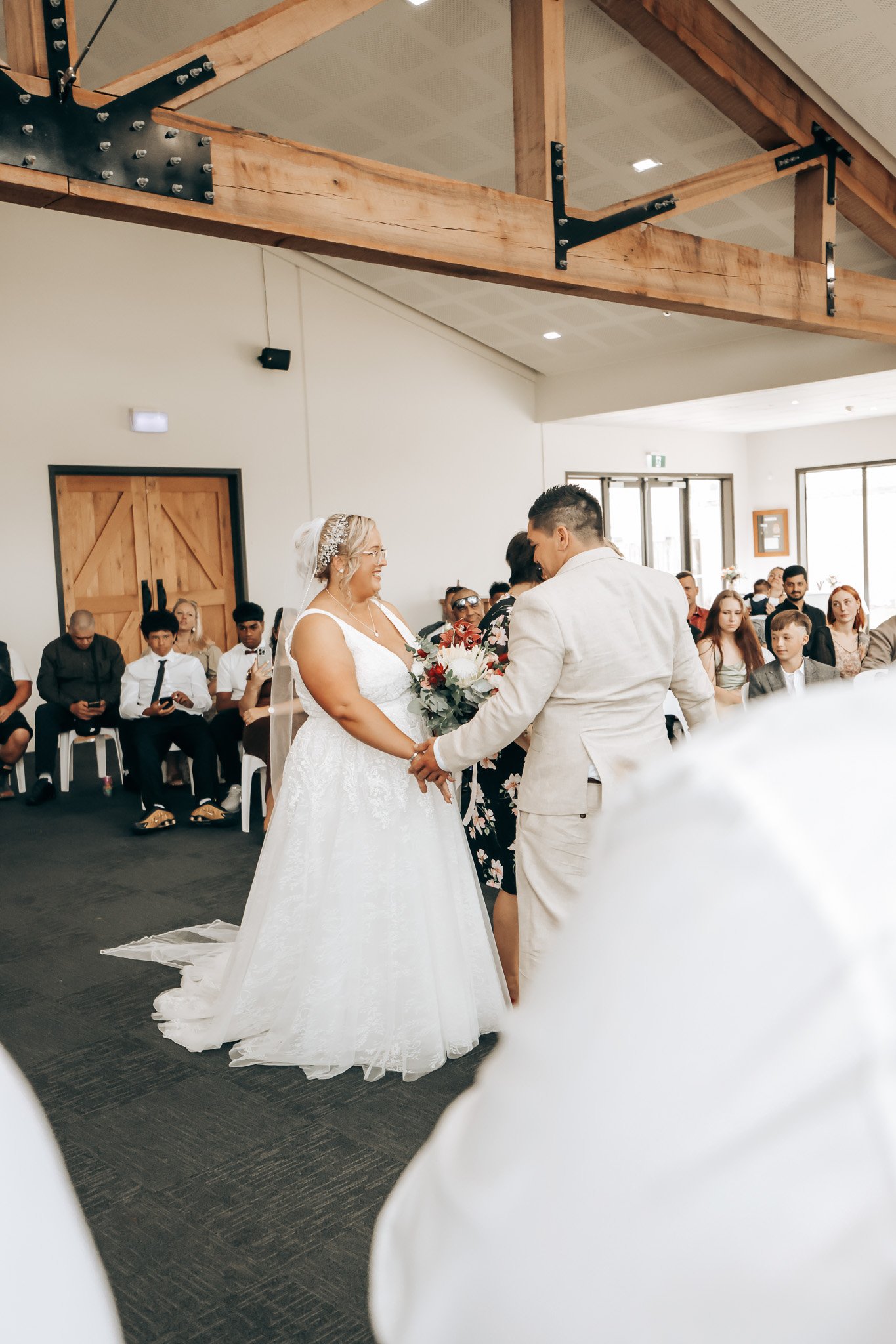 Wedding ceremony with a bride and groom holding hands in a decorated indoor venue, surrounded by seated guests. The bride wears a white wedding gown and the groom a light-colored suit.