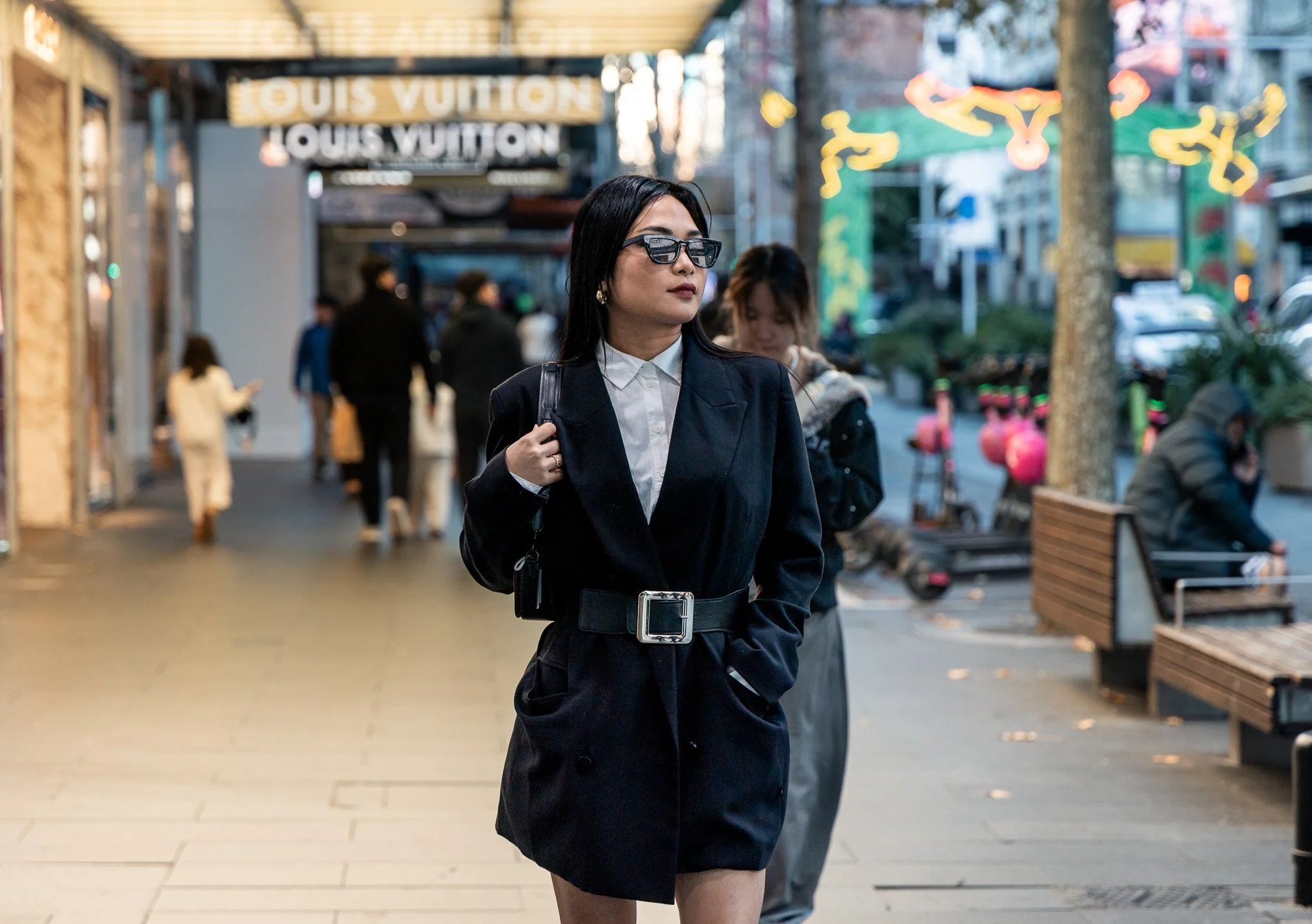 A woman with black hair, sunglasses, and earrings wearing a black blazer, white shirt, and black skirt, walking outdoors in an urban setting with shops, people, and colorful decorations.