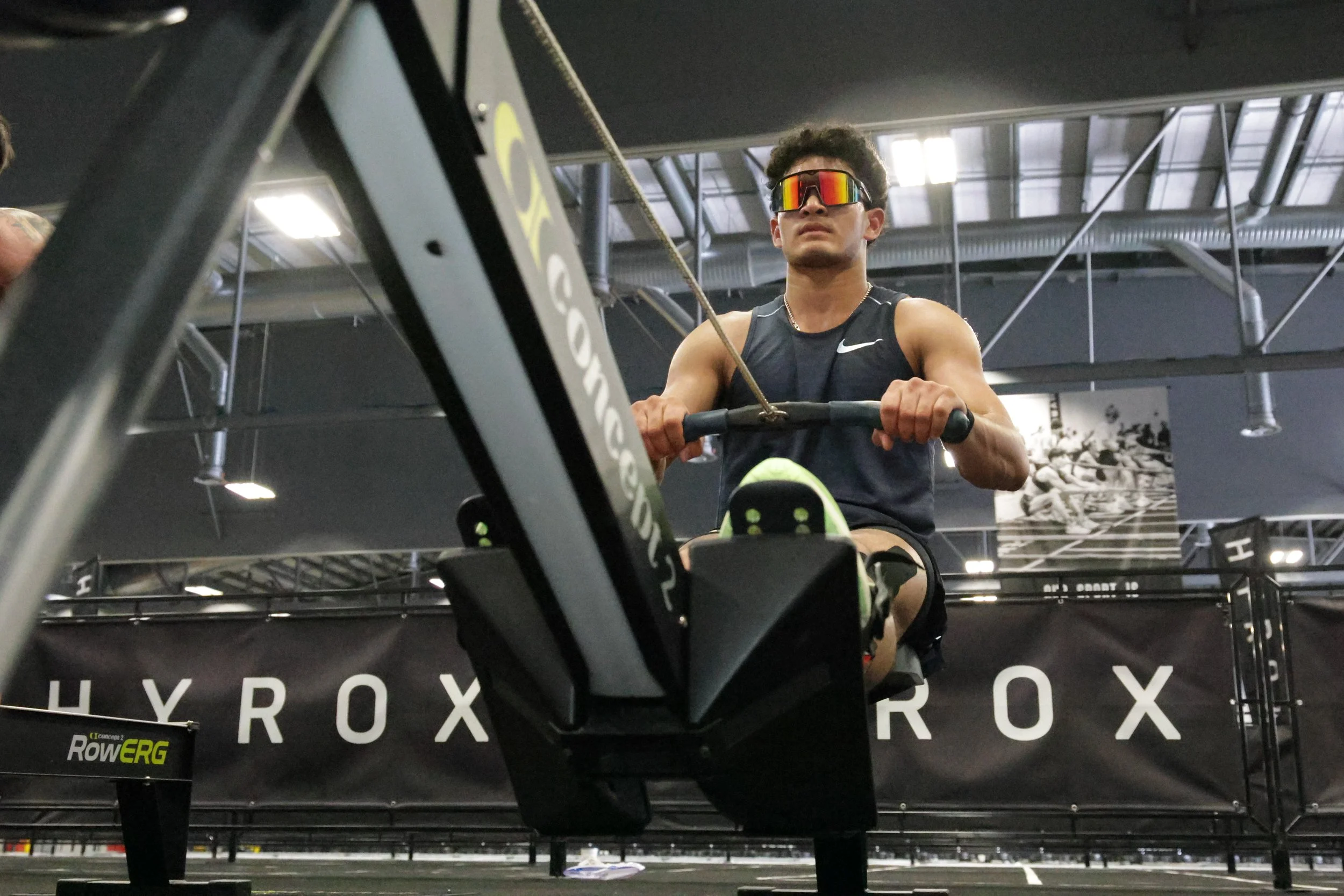 A man in a sleeveless black athletic shirt and reflective sunglasses is using a rowing machine in a gym.