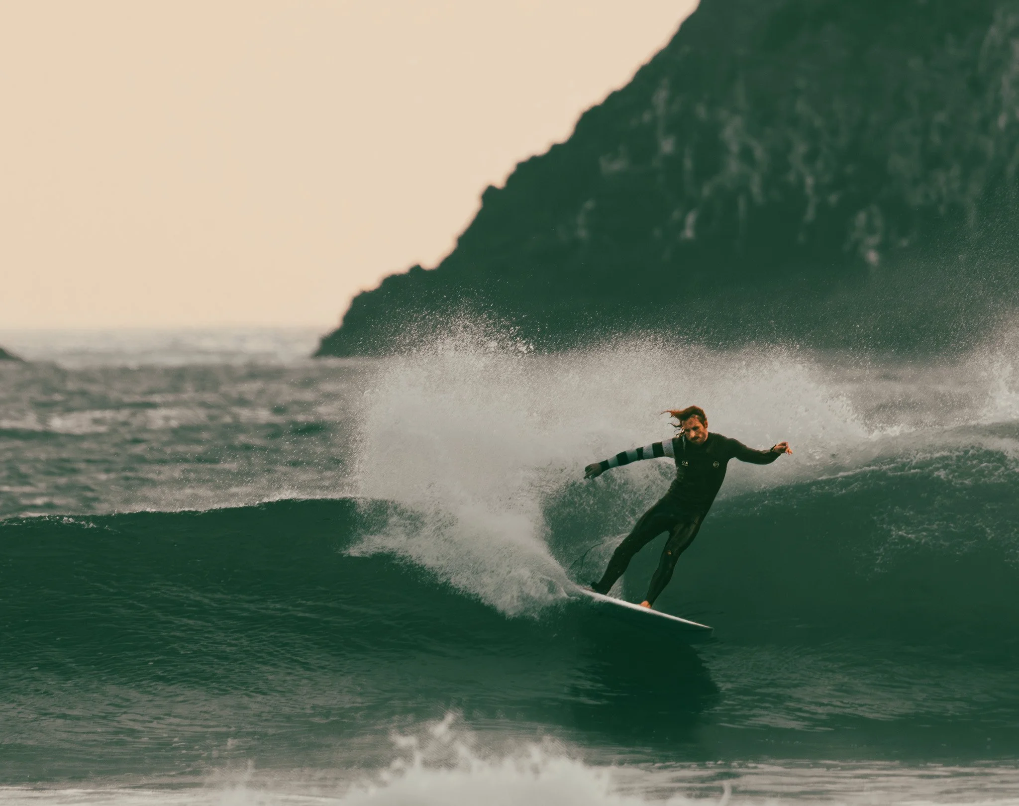 A person snowboarding on a wave in the ocean, with a large rock formation in the background.