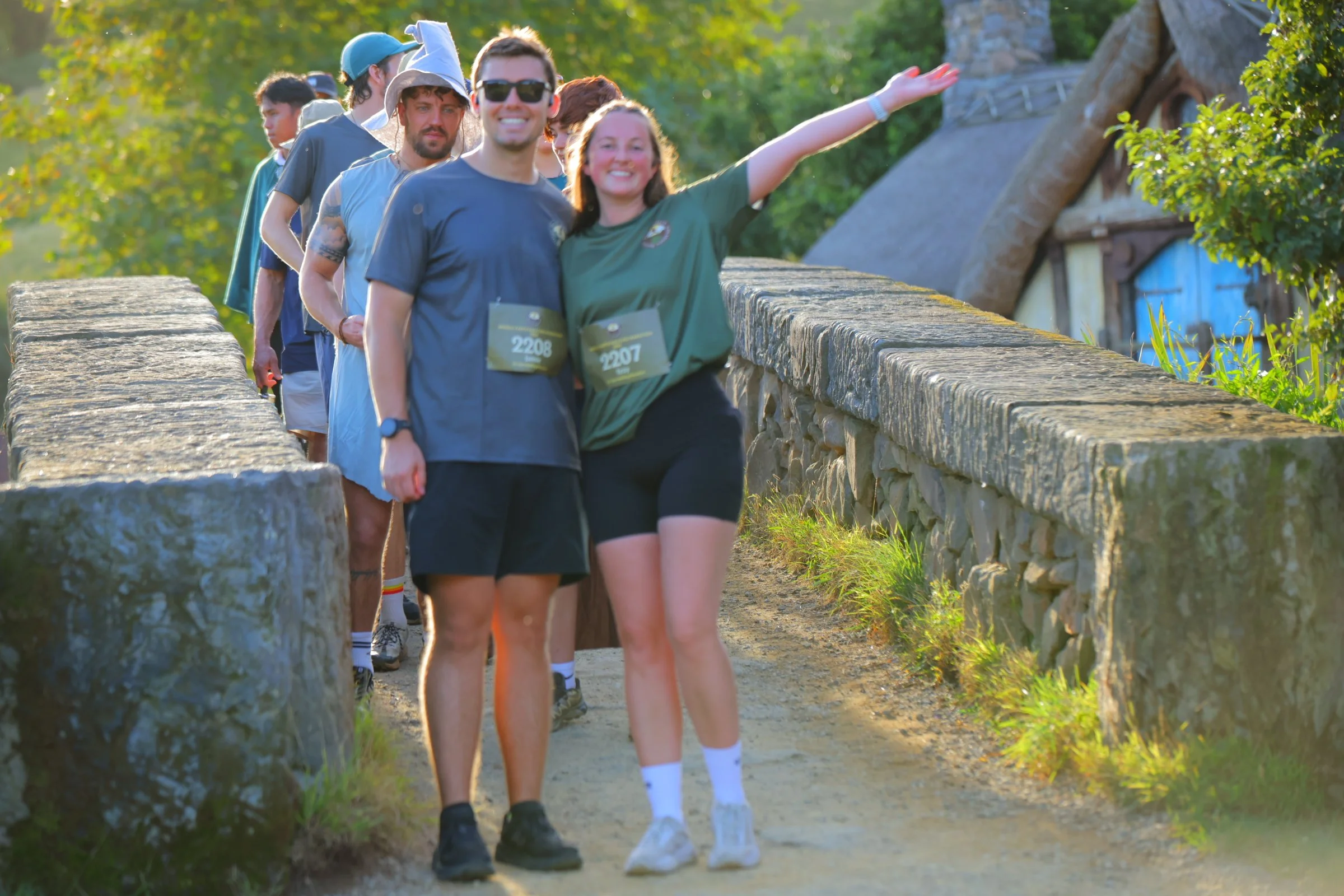 A group of people walking on a stone path outdoors, with one woman in front and smiling, dressed in a green shirt and black shorts. The others are behind her, some wearing sunglasses and casual clothing, with a sunny, green, leafy background.
