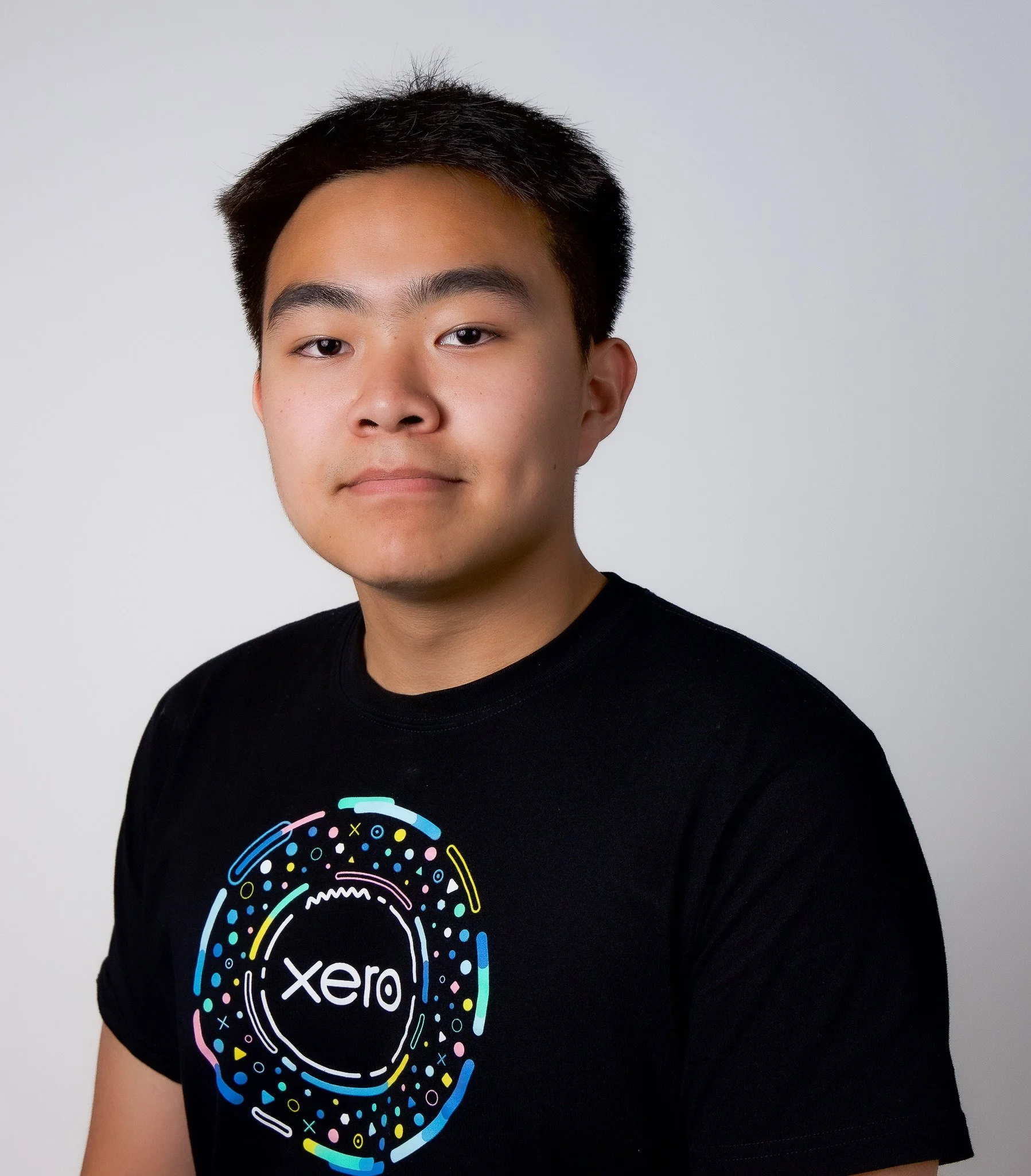A young man with short black hair and a slight smile wearing a black t-shirt with a colorful circular design and the word 'xero' on it, standing against a plain white background.