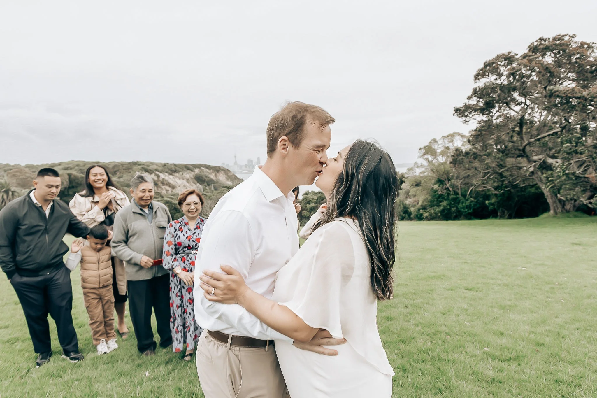 A couple sharing a kiss while holding each other outdoors in a park on a cloudy day, with friends and family smiling and watching in the background.