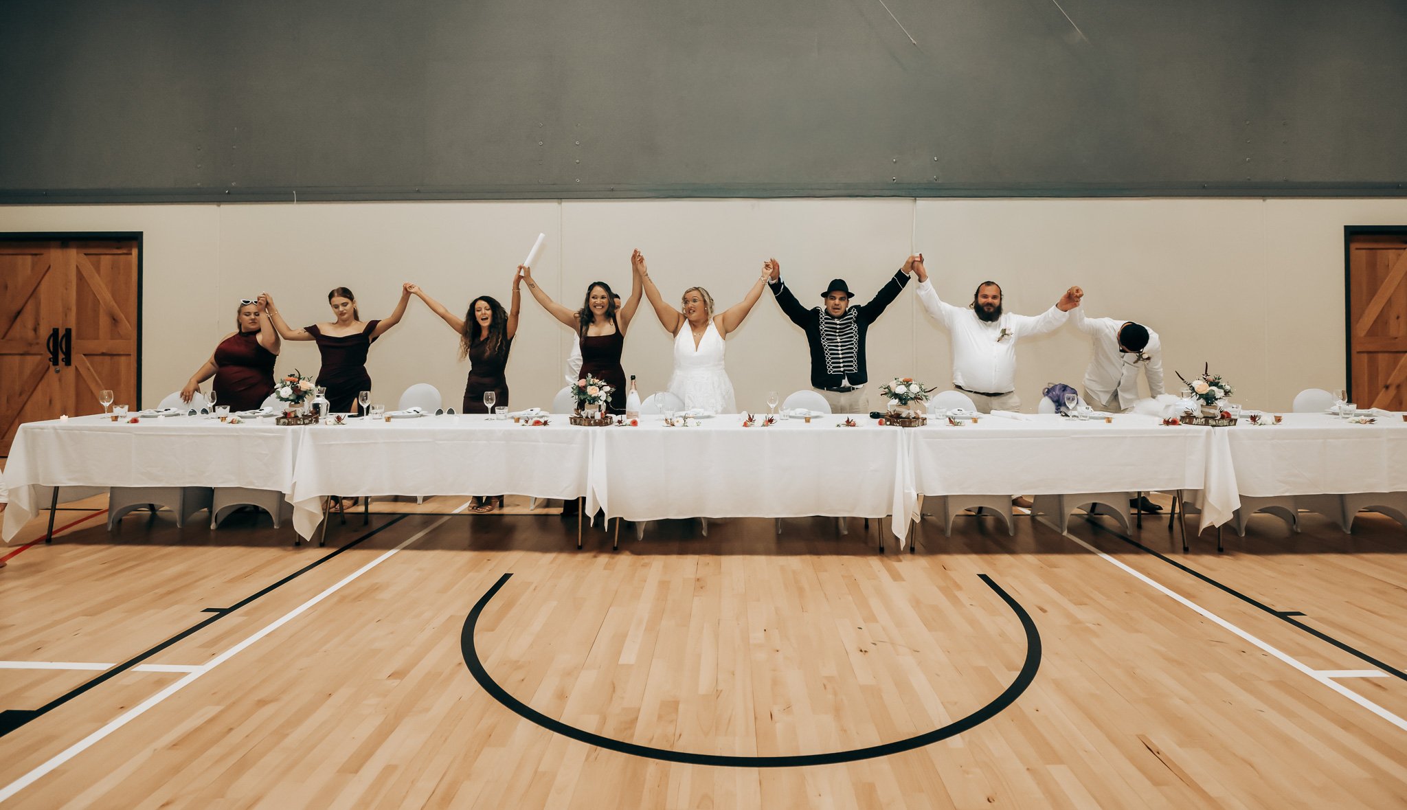 People standing behind a long decorated table at a wedding reception, holding hands and raising their arms, in a gymnasium with wooden floors and closed wooden doors.