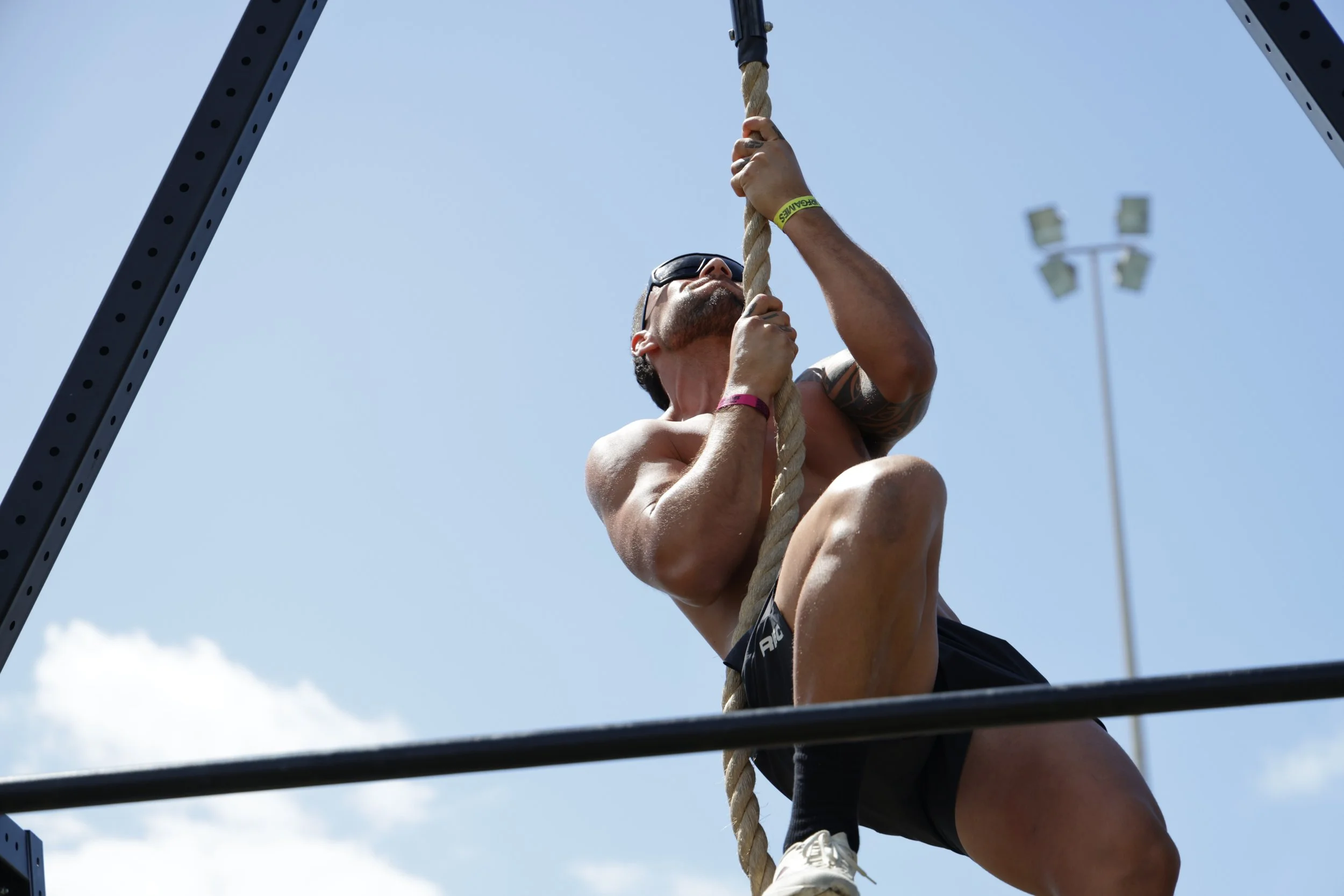 A man wearing sunglasses and a yellow wristband climbs a thick rope during an outdoor fitness event, with a sports field light in the background and a clear blue sky.