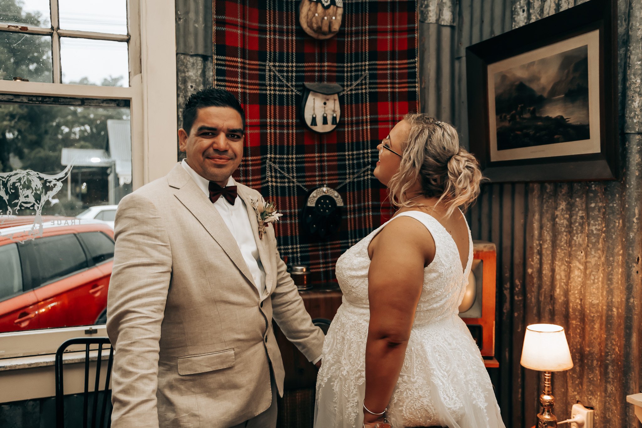 A man in a beige suit with a white shirt and bow tie, and a woman in a white lace wedding dress with glasses, standing indoors in front of a red plaid wall hanging, near a window and a framed landscape painting, a lit lamp on a side table, and a red 