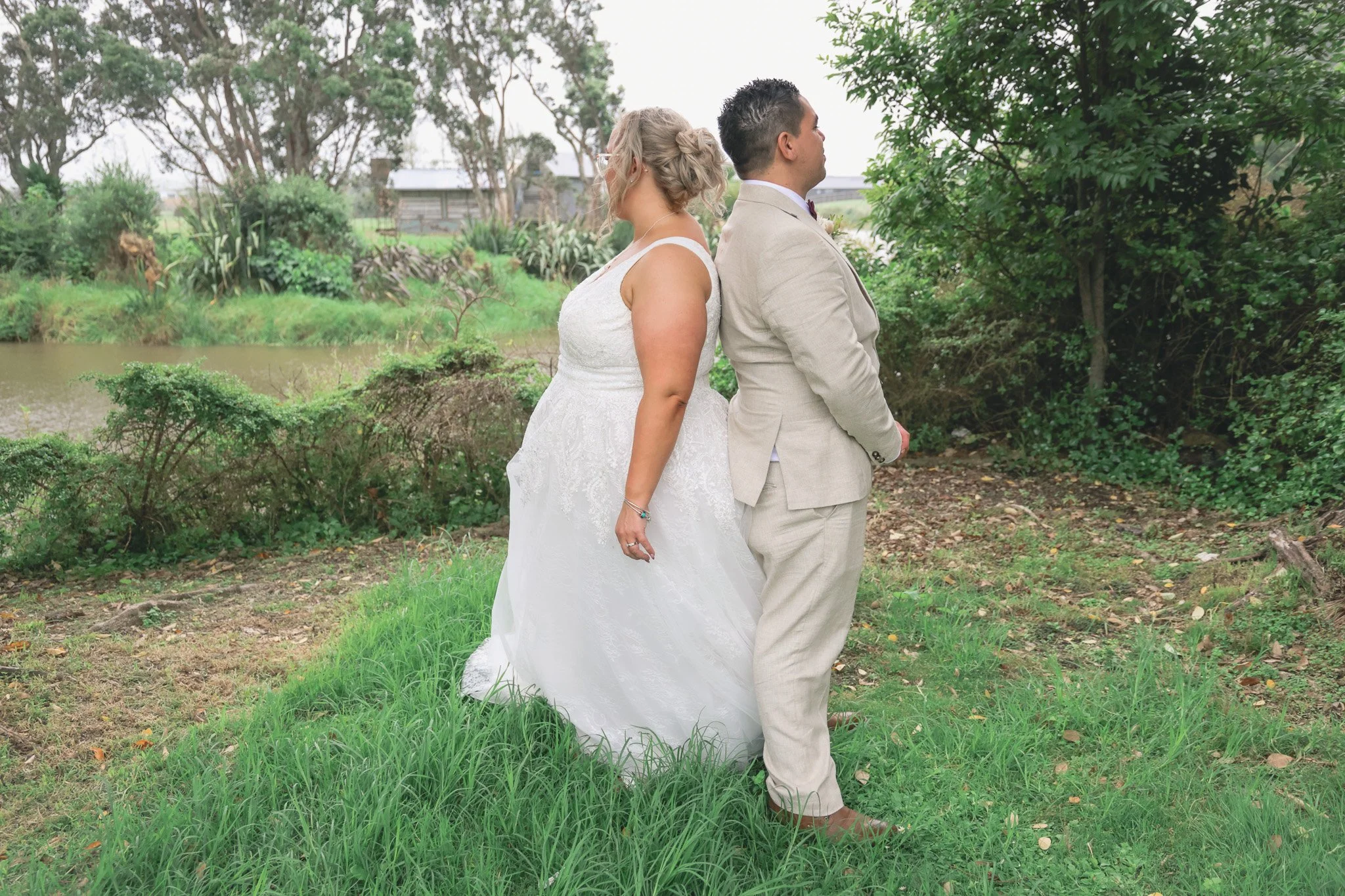 A bride and groom stand back to back outdoors near a pond, surrounded by greenery and trees, during a wedding ceremony.