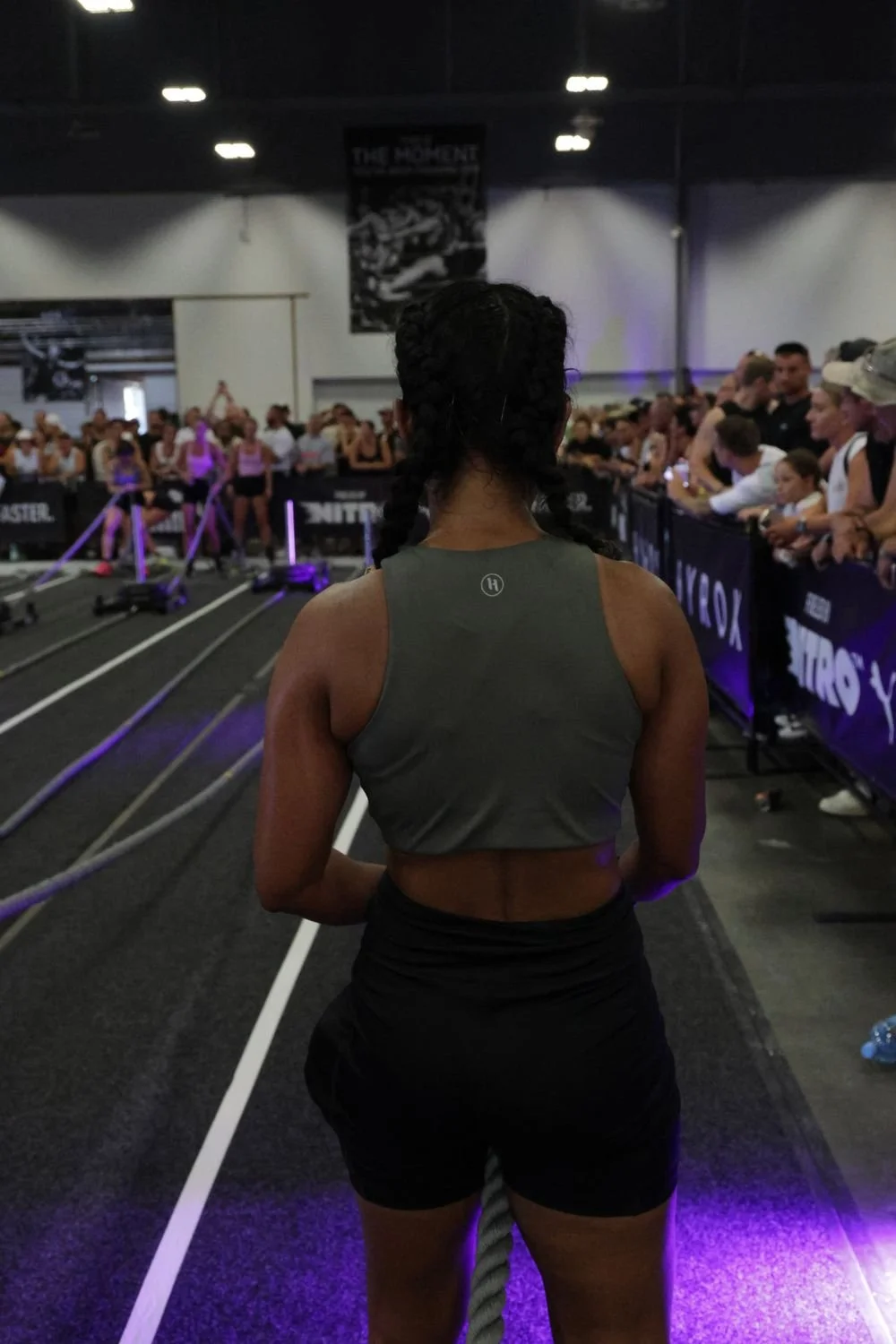 A woman with braided hair standing at the start line of a CrossFit competition, with a large crowd of spectators behind her and workout rigs on the floor.