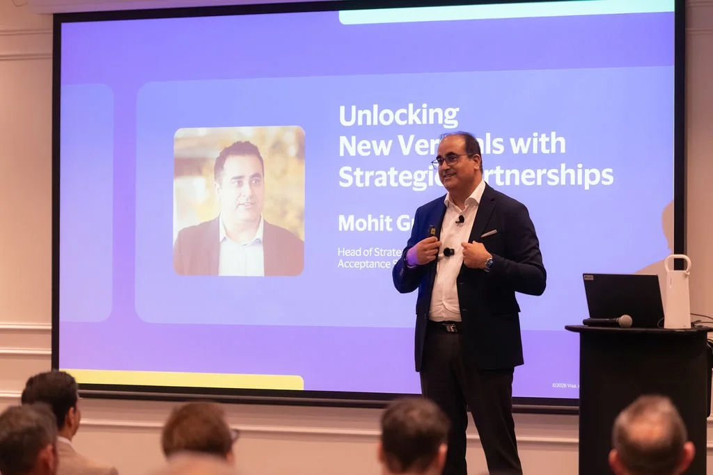 A man in a suit standing on stage giving a presentation to an audience. The presentation slide behind him reads "Unlocking New Verticals with Strategic Partnerships" and features a photo of another man and his title, "Mohit G... Head of Strategy Acce