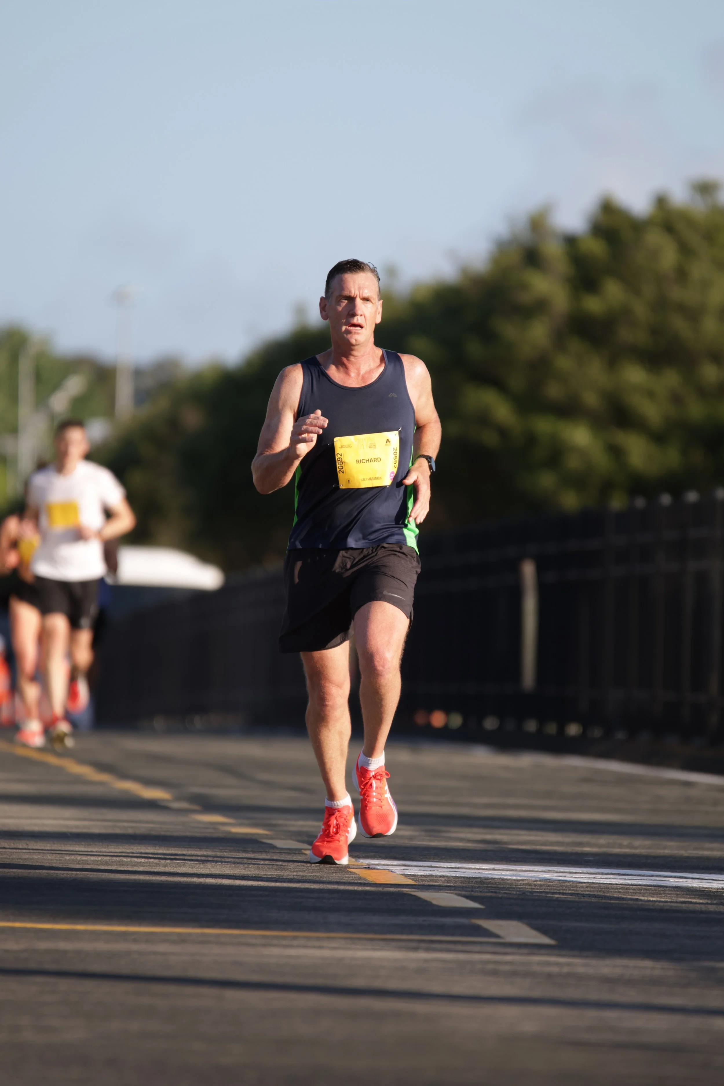 A man running in a marathon wearing a navy tank top, black shorts, and red running shoes, with others in the background, on a paved road lined with trees.