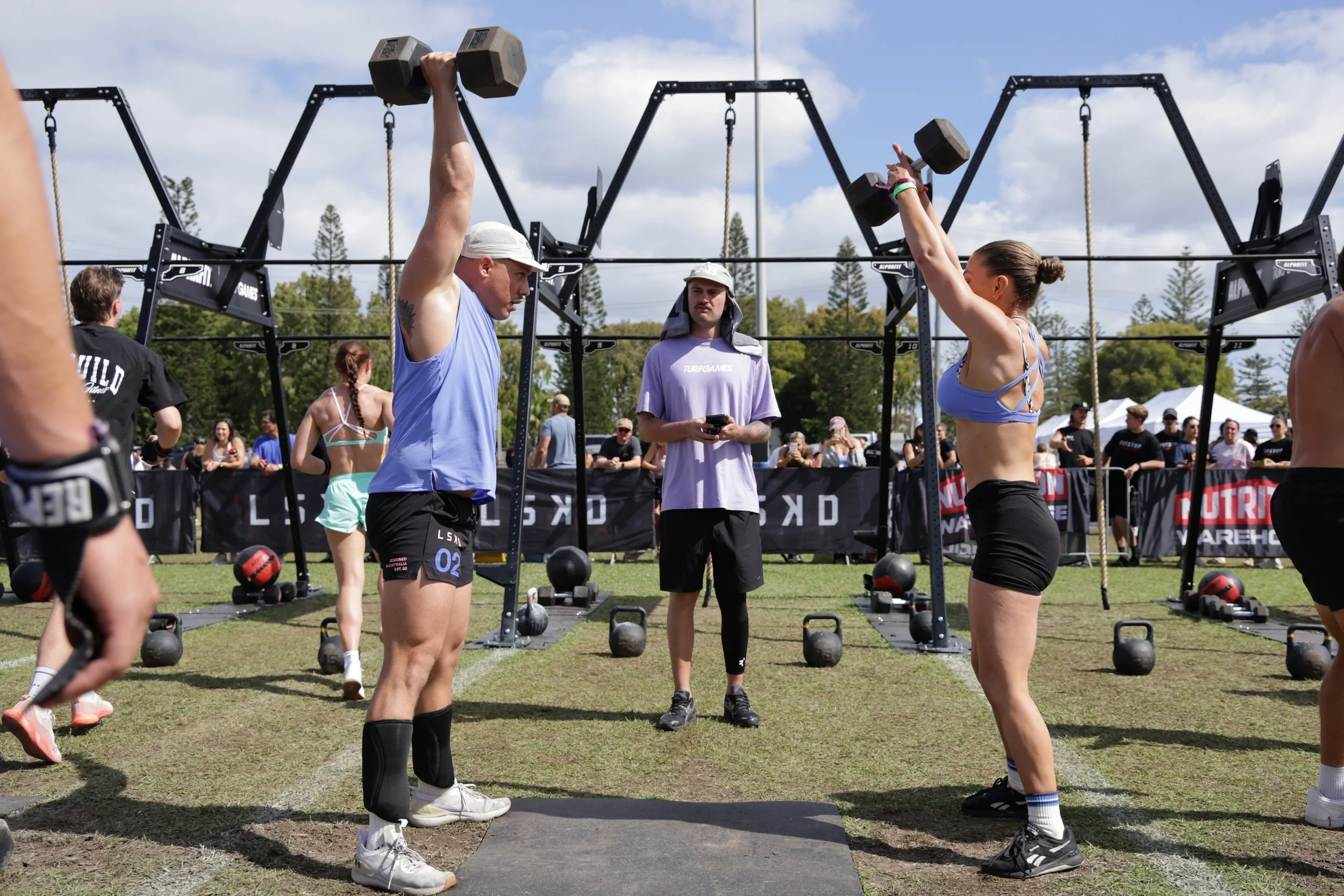 People participating in an outdoor fitness competition or event, lifting dumbbells and engaging in strength training exercises with a fitness station setup under a partly cloudy sky.