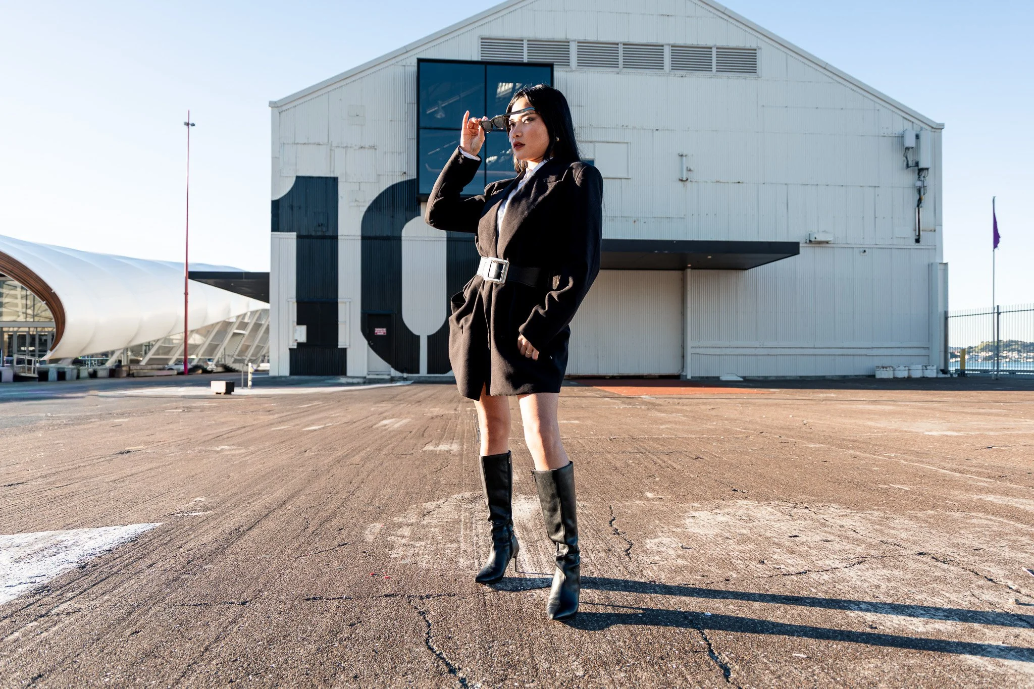 A woman in black boots and a stylish black coat stands outdoors with a large building in the background, adjusting her glasses as she looks into the distance.