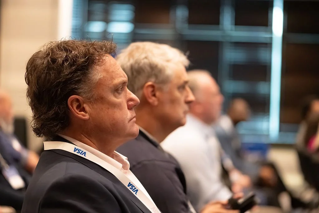 Side profile of a man with curly brown hair wearing a suit and an ID badge that says VISA, attending a conference or seminar with other attendees.