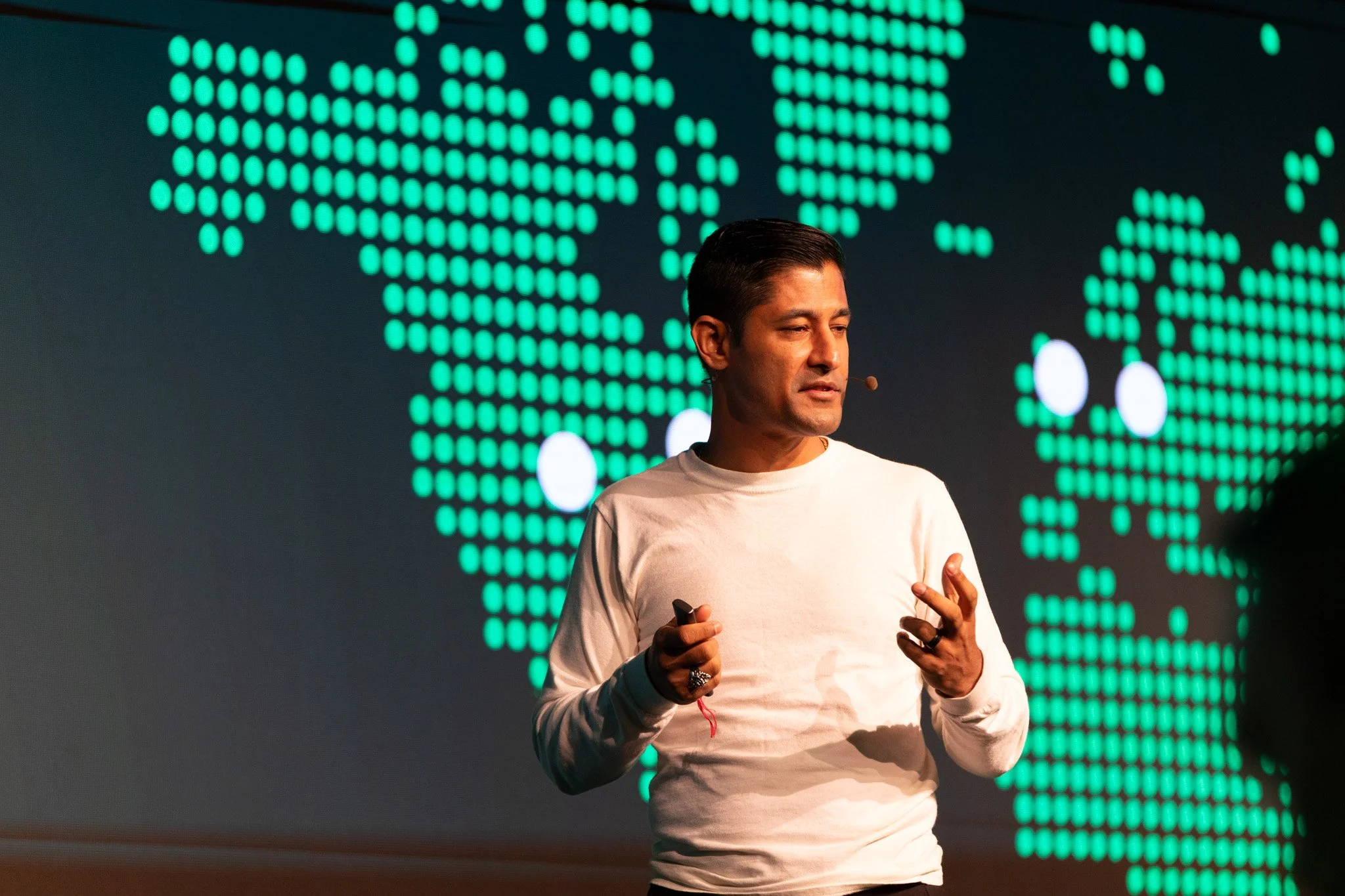 A man wearing a white long sleeve shirt and a microphone headset giving a presentation on a stage with a large digital world map displayed behind him.