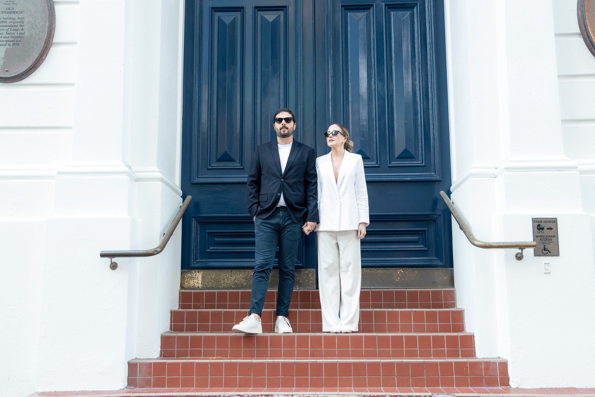 A man and woman dressed in formal attire standing on red brick steps in front of a large, dark blue door with white framing. Both are wearing sunglasses, and the man has a beard and mustache, while the woman has her hair tied back. The woman is holdi